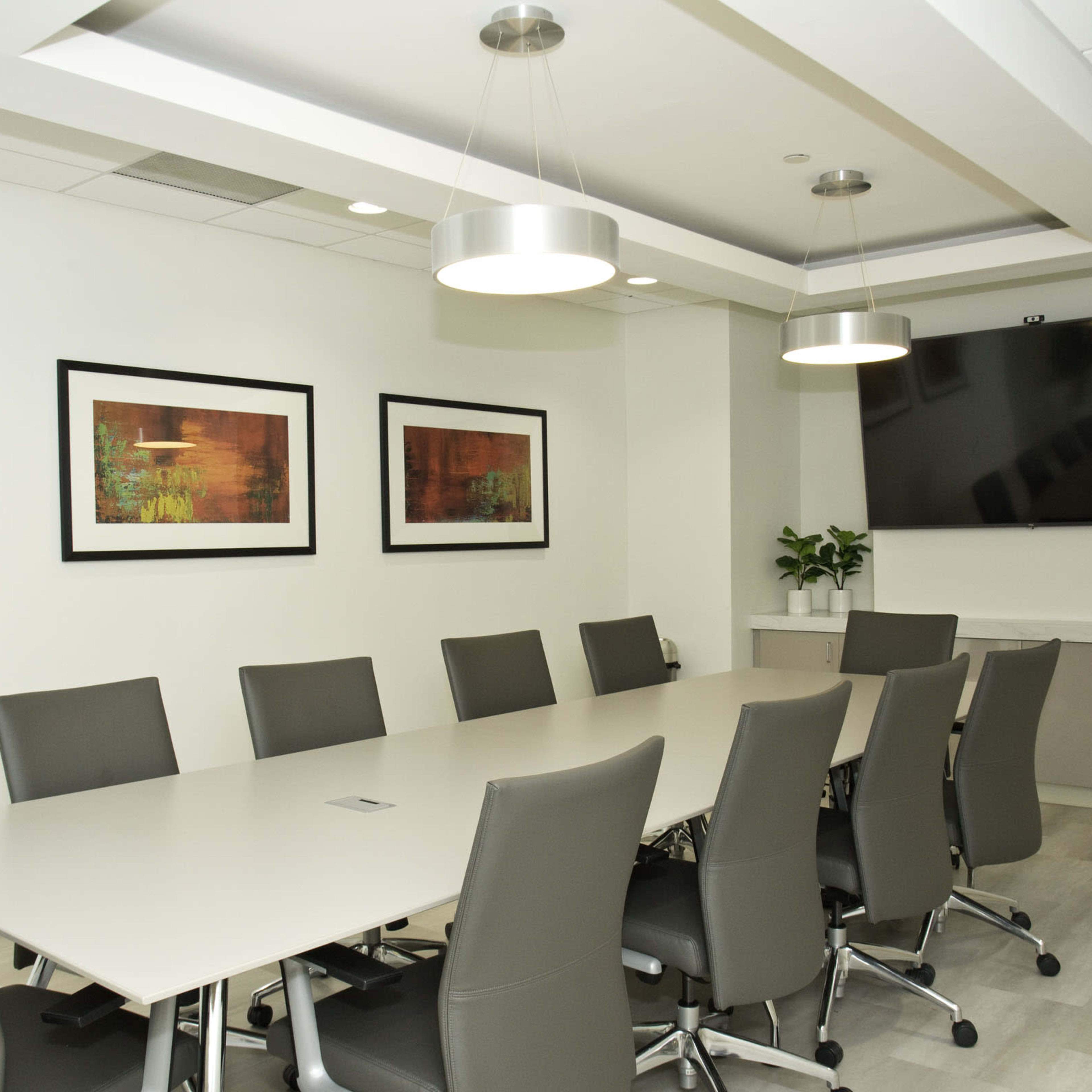 A conference room features a long table surrounded by gray chairs, with framed artwork on the walls and a television mounted in one corner.