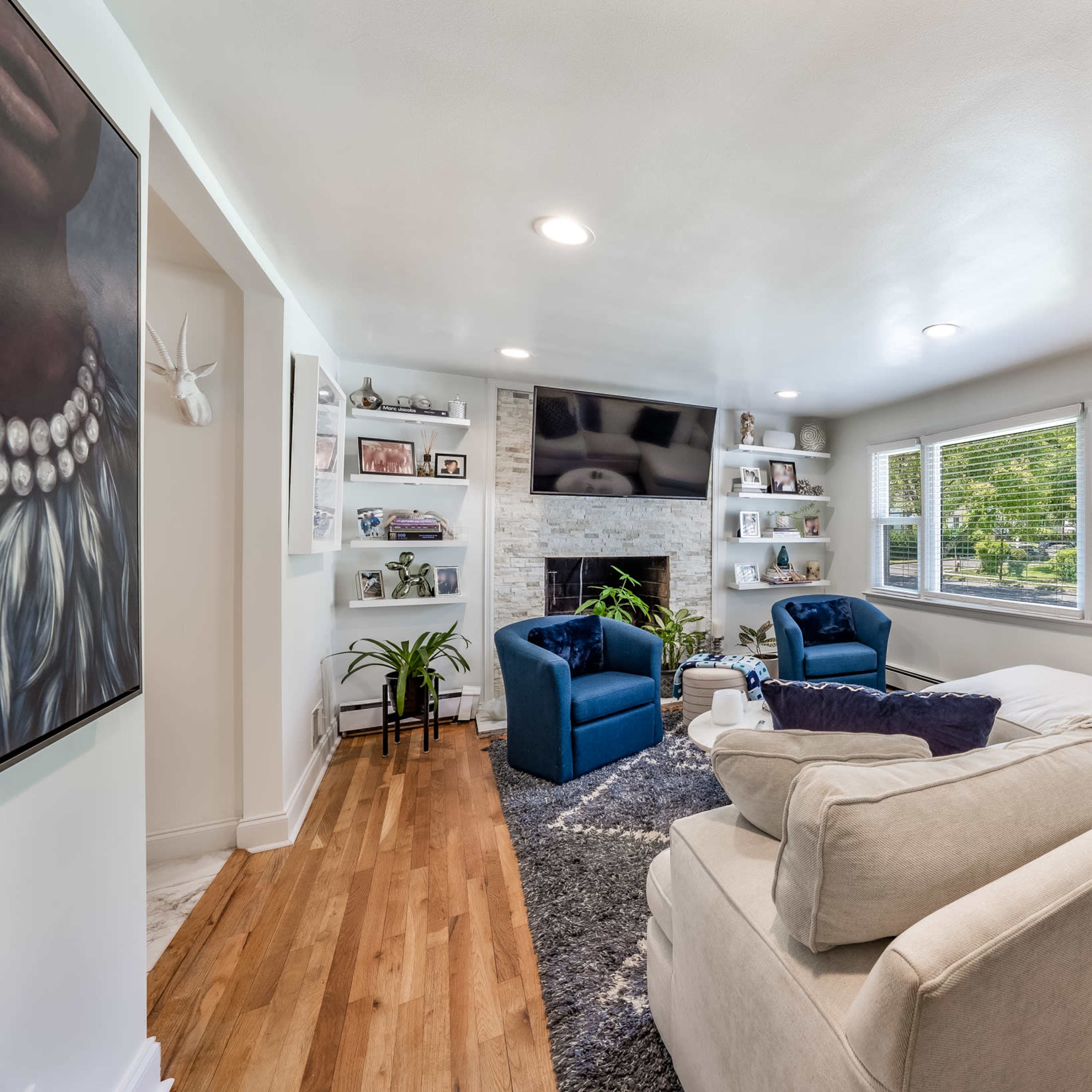 A living room features a light-colored couch, two blue chairs, a fireplace with a television above it, and shelves filled with photographs and plants.