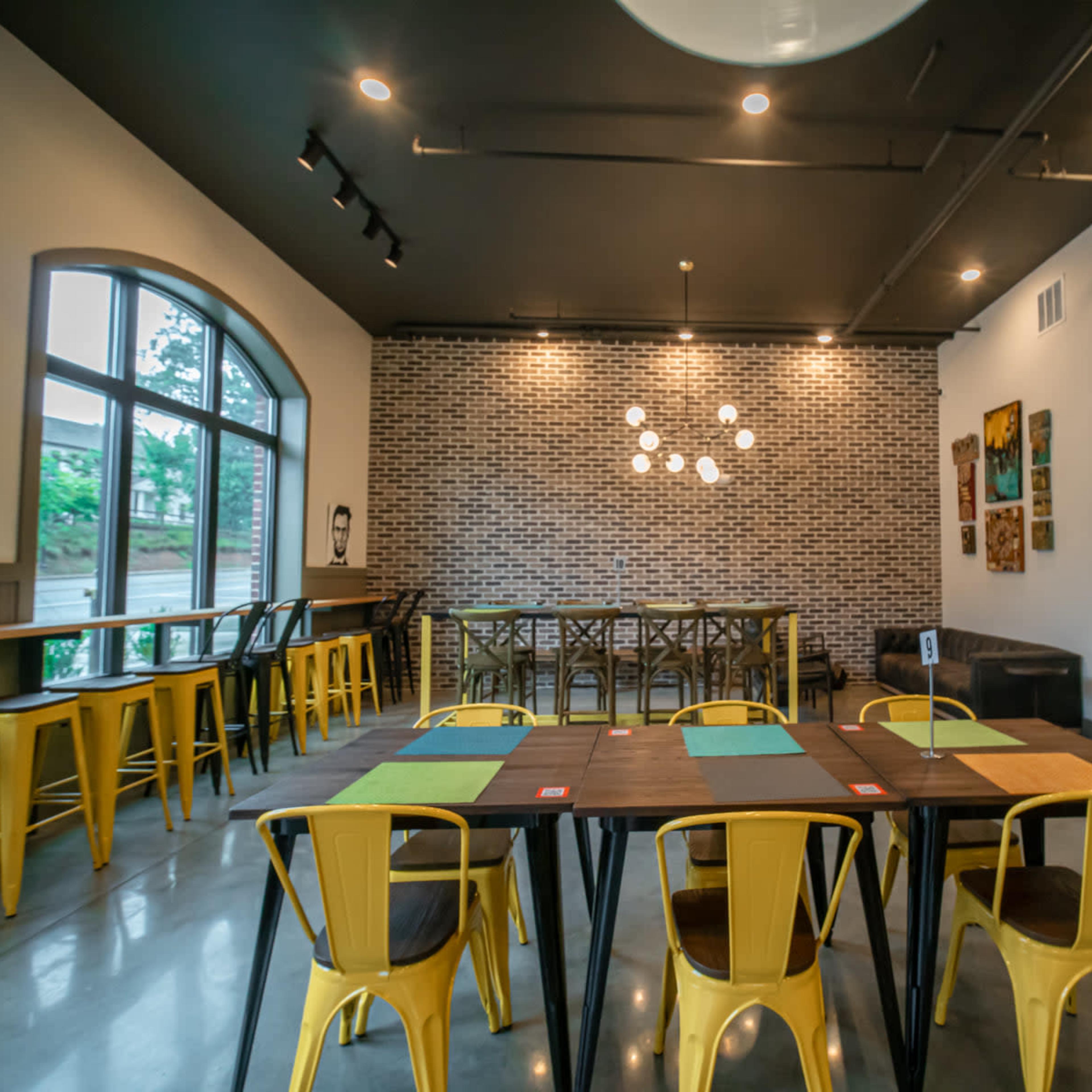 A modern dining space featuring yellow chairs, a large wooden table, and a brick accent wall with pendant lights overhead.