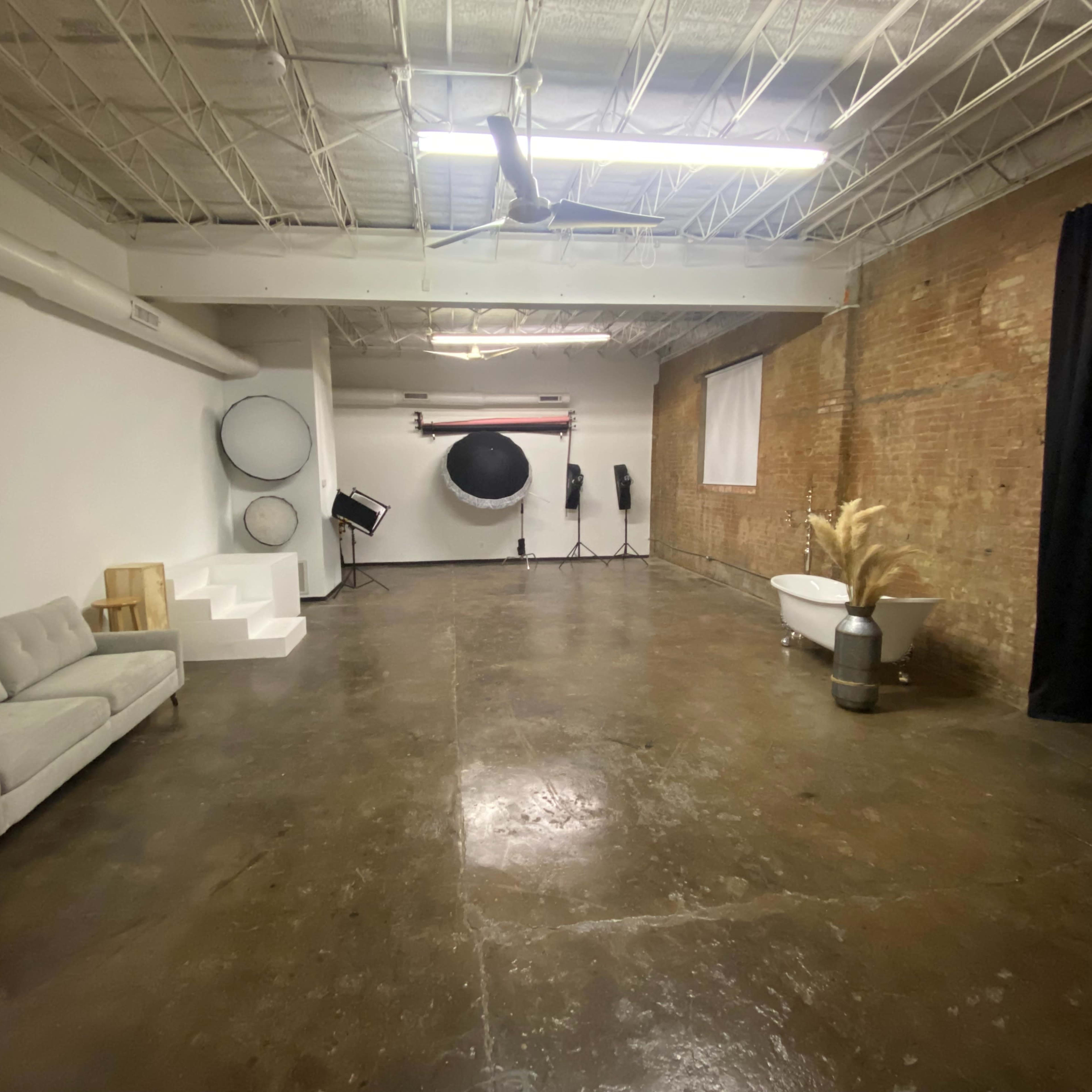A spacious, industrial-style studio featuring a gray couch, a circular backdrop, and various photography equipment against a brick wall.