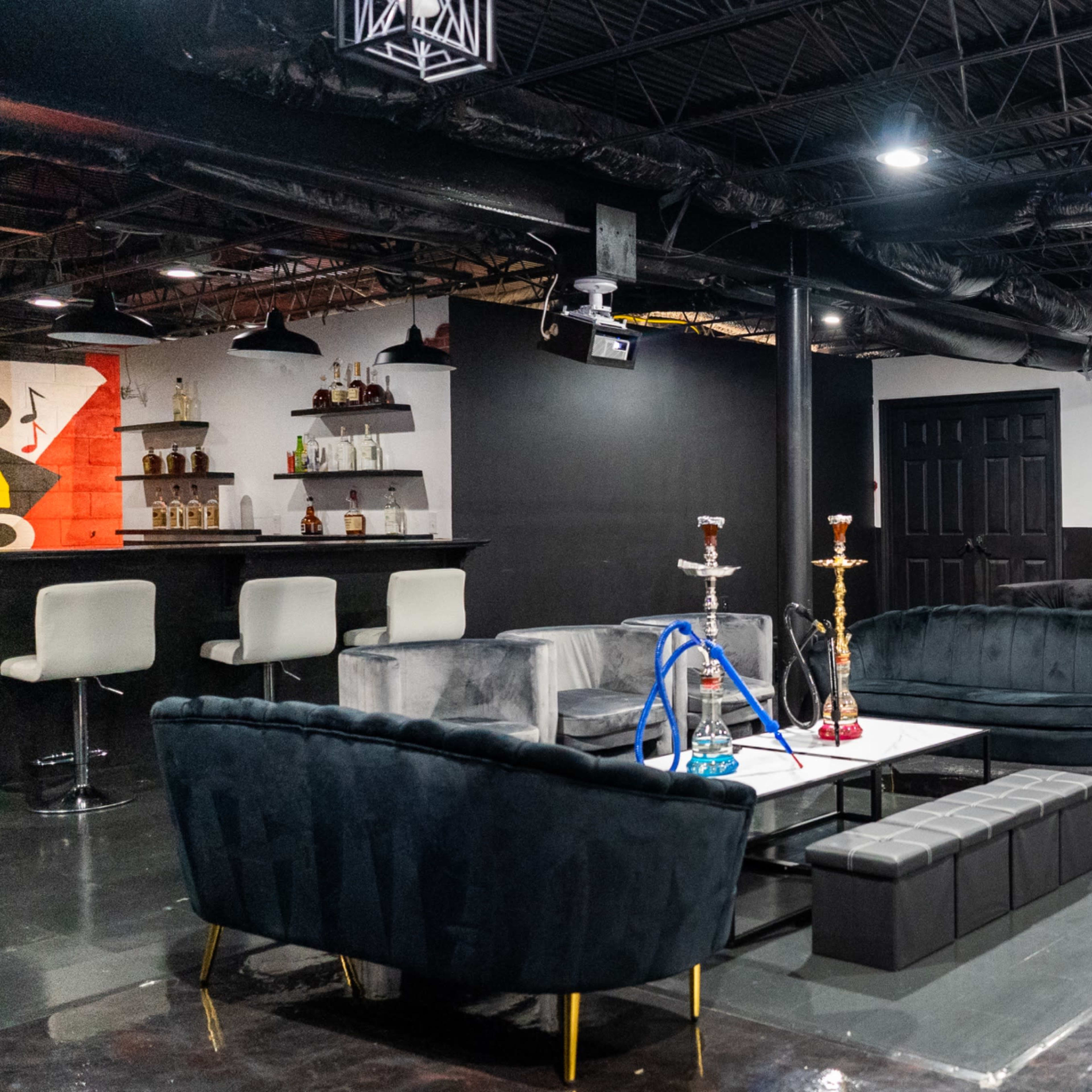 A spacious lounge area featuring dark furniture, a bar with liquor bottles, and a setup for hookahs in a modern interior.