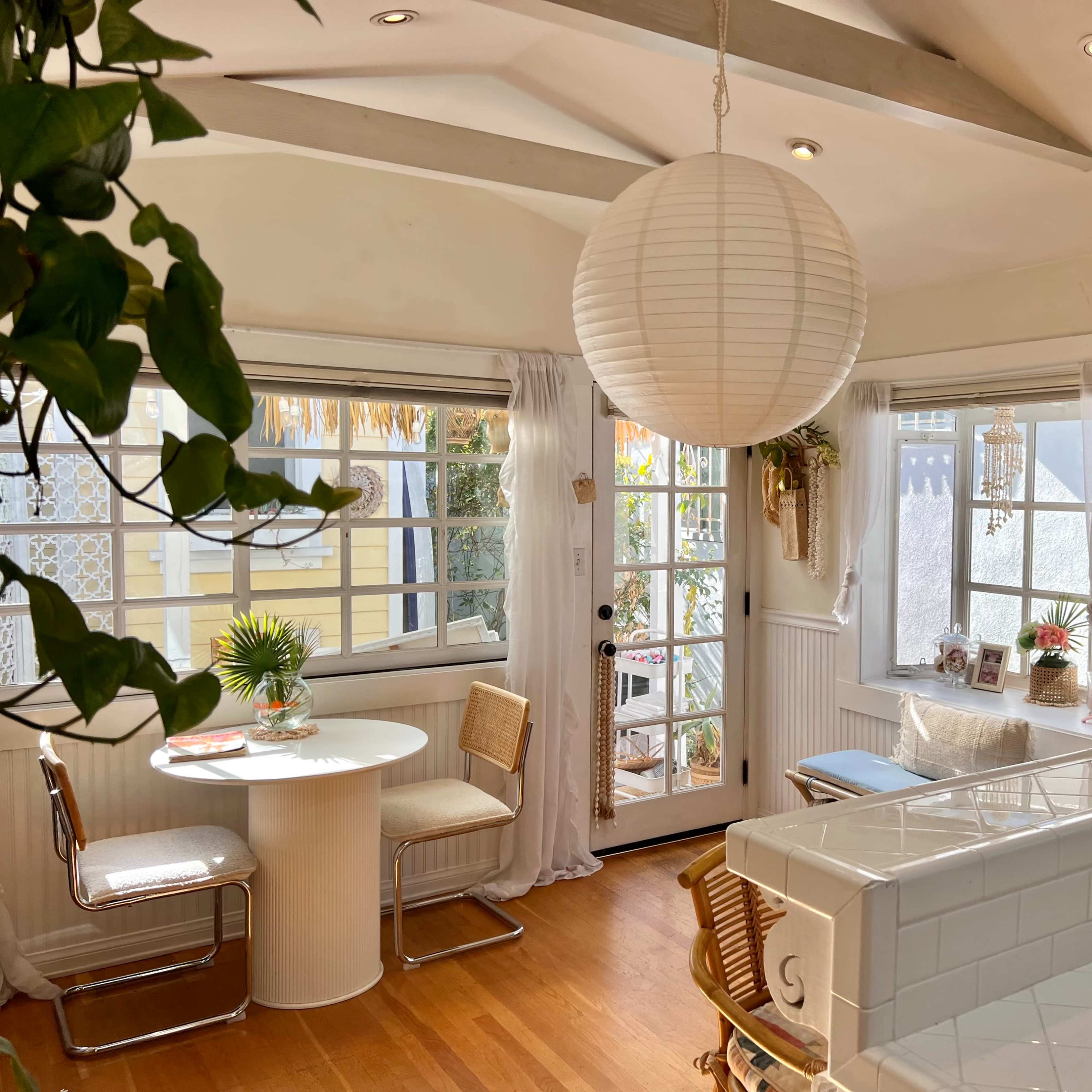 A bright dining area features a white round table with two chairs, large windows, and a paper lantern hanging from the ceiling.