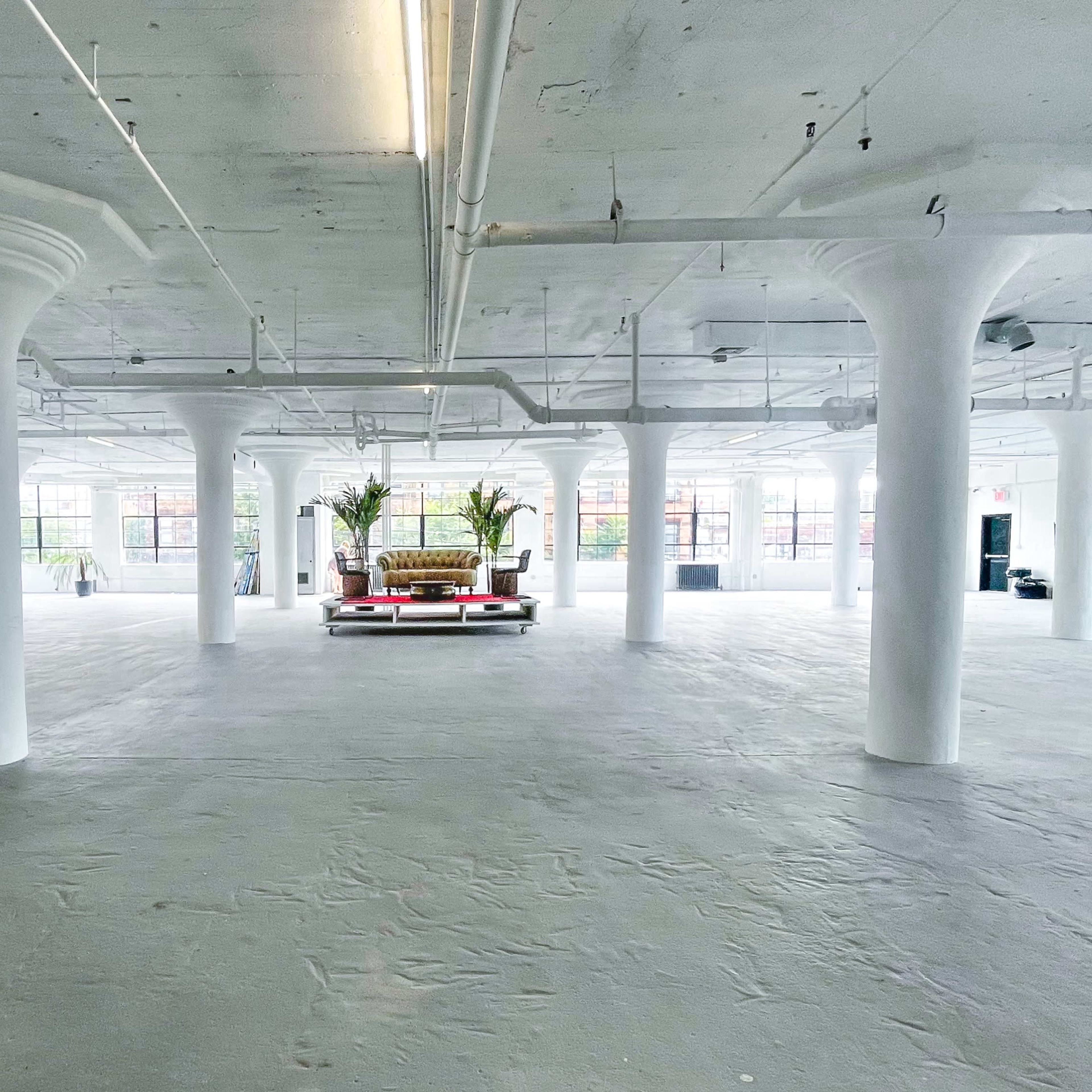A spacious, empty room with large windows, white walls, and several columns, featuring a sofa at the center.