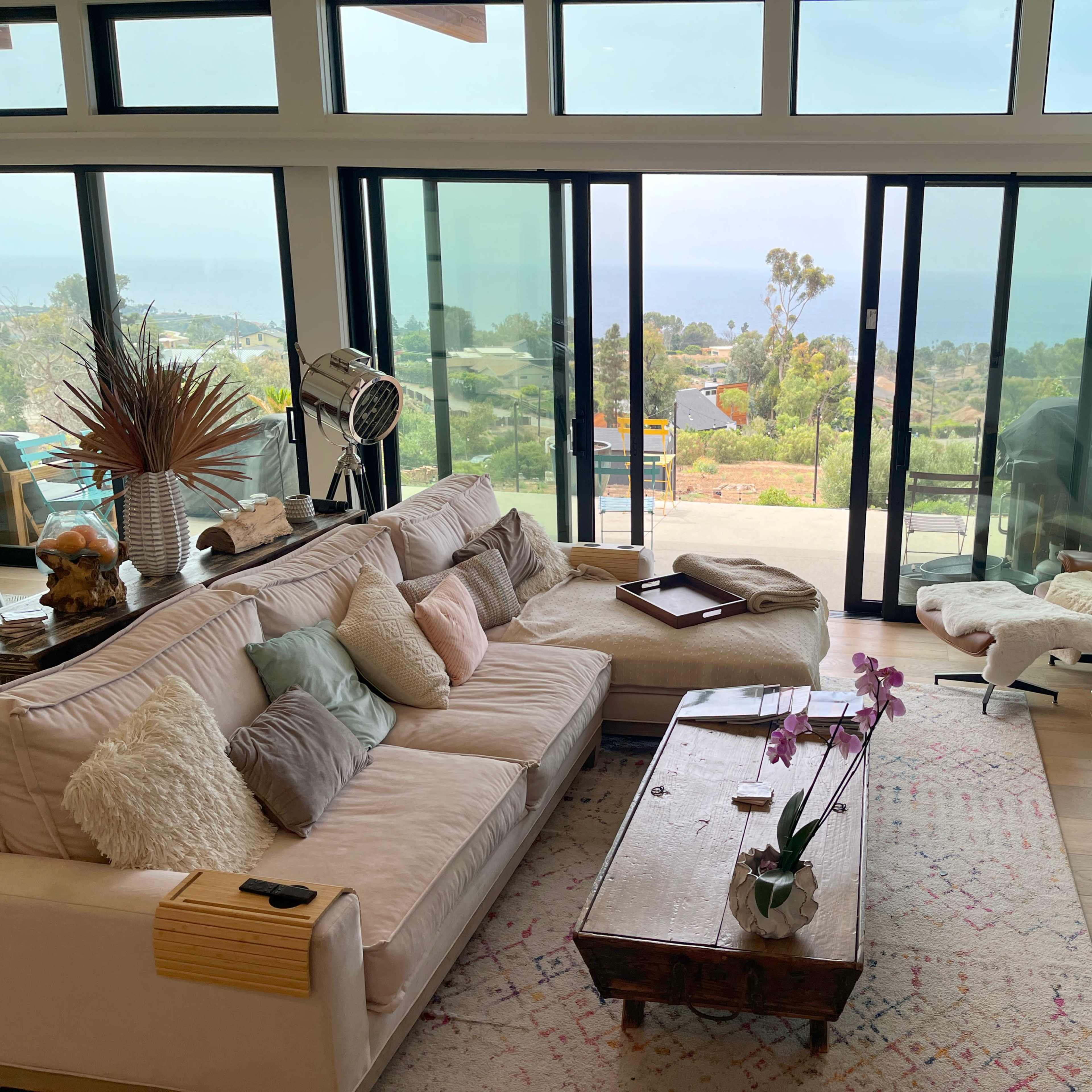 A modern living room with large windows overlooking a coastal view, featuring a sectional sofa, a wooden coffee table, and a lounge chair.