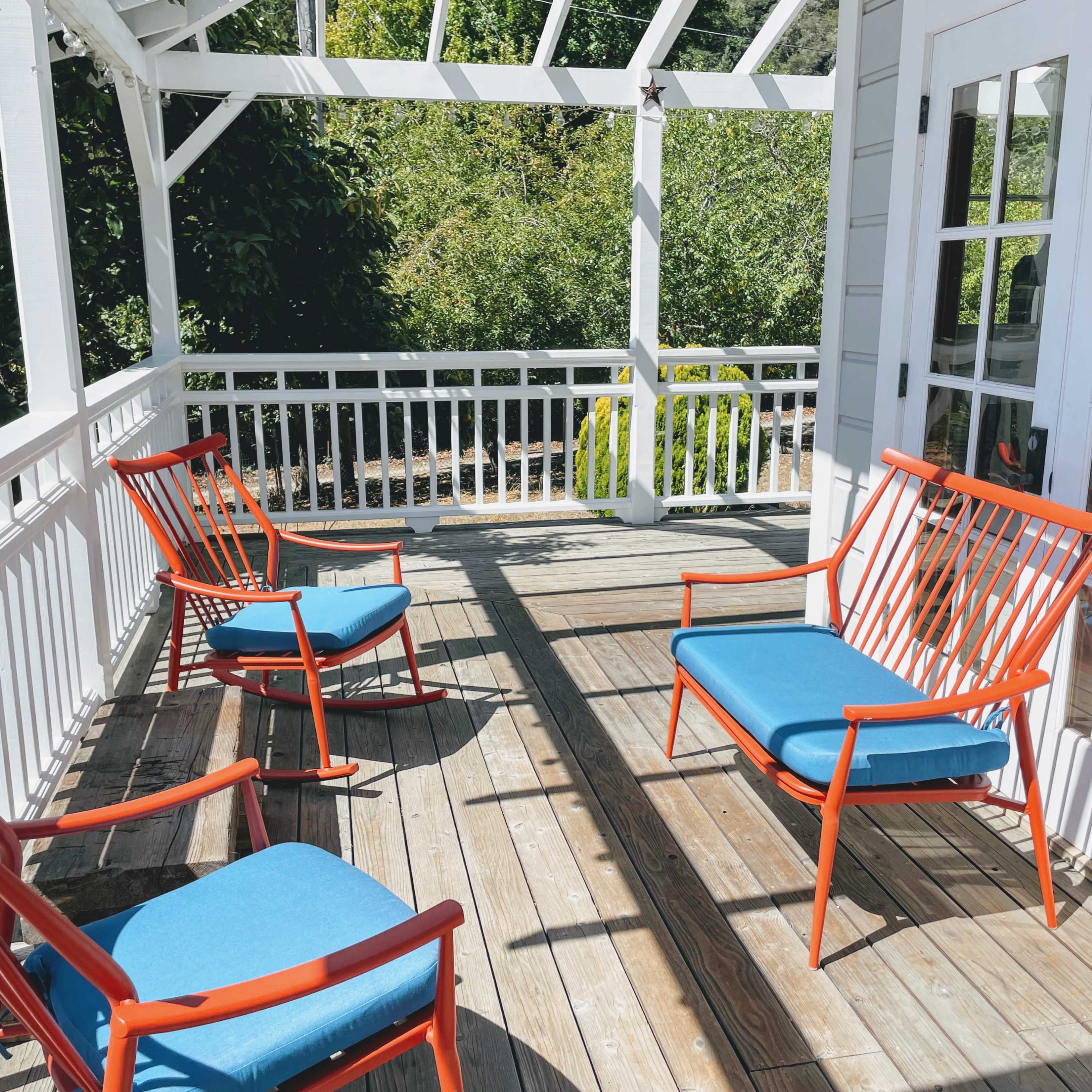 A bright porch features four chairs with blue cushions and orange frames arranged around a wooden deck.