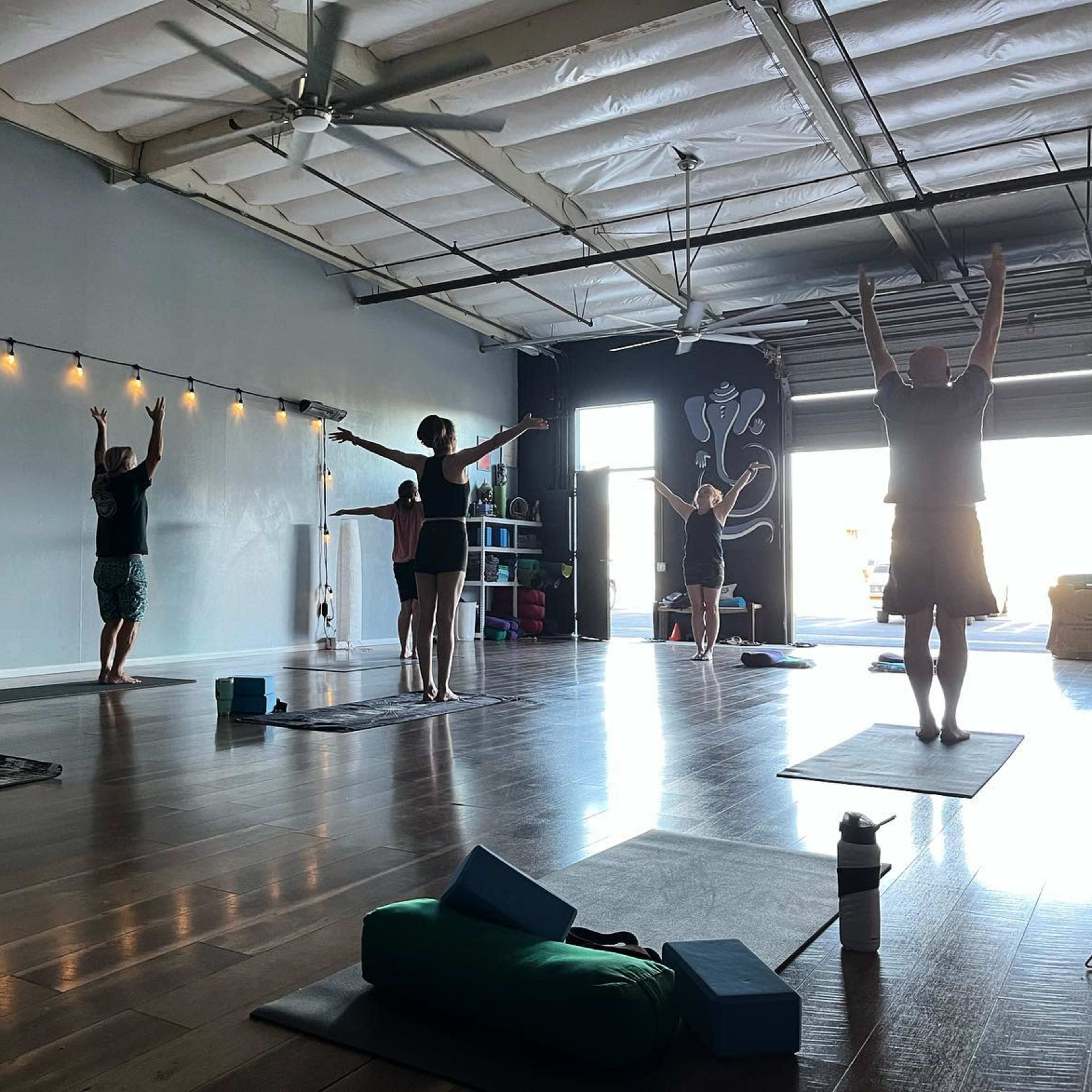 A group of six people is practicing yoga in a spacious studio with wooden flooring and large windows.
