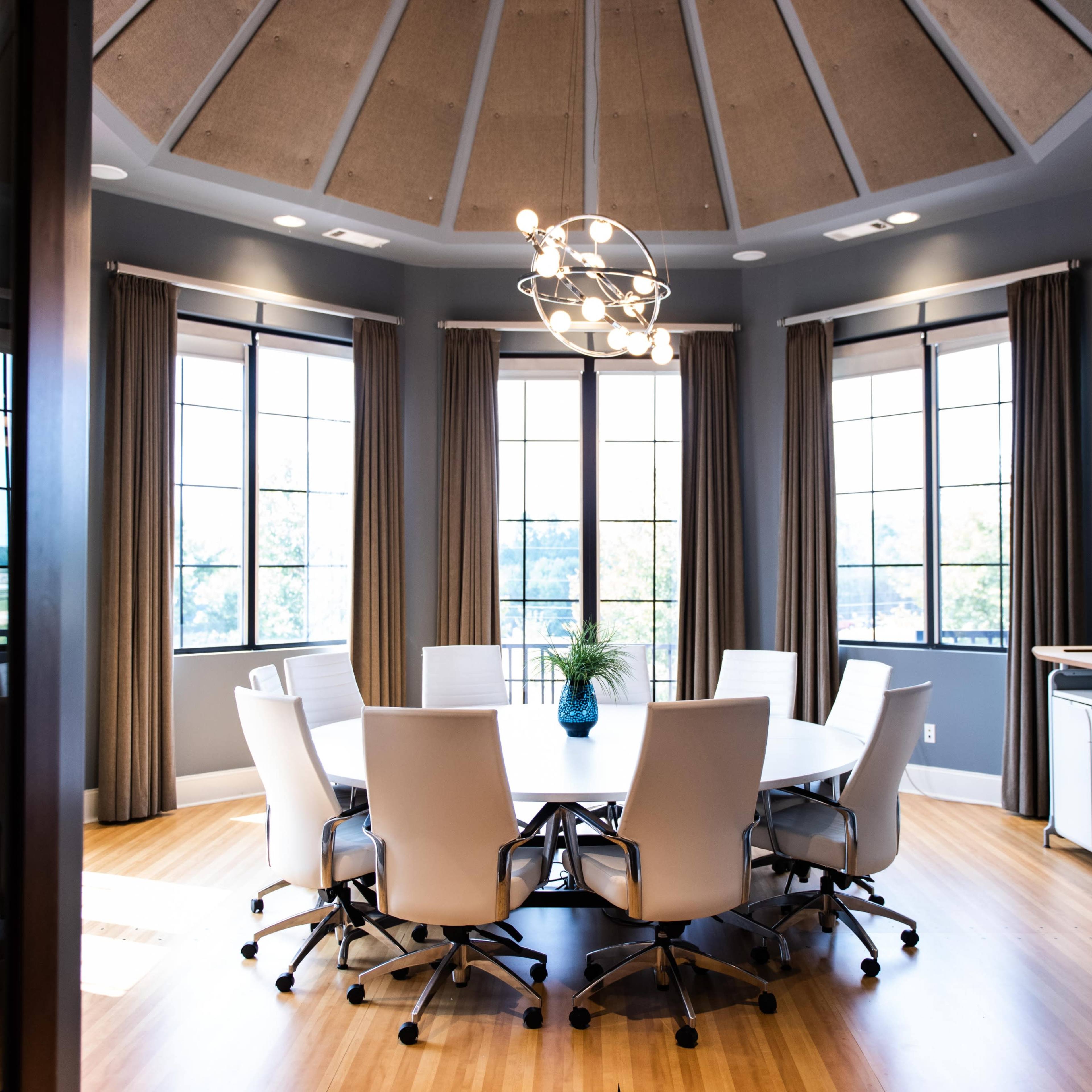 A modern conference room features a round table surrounded by white chairs, large windows, and a central light fixture.