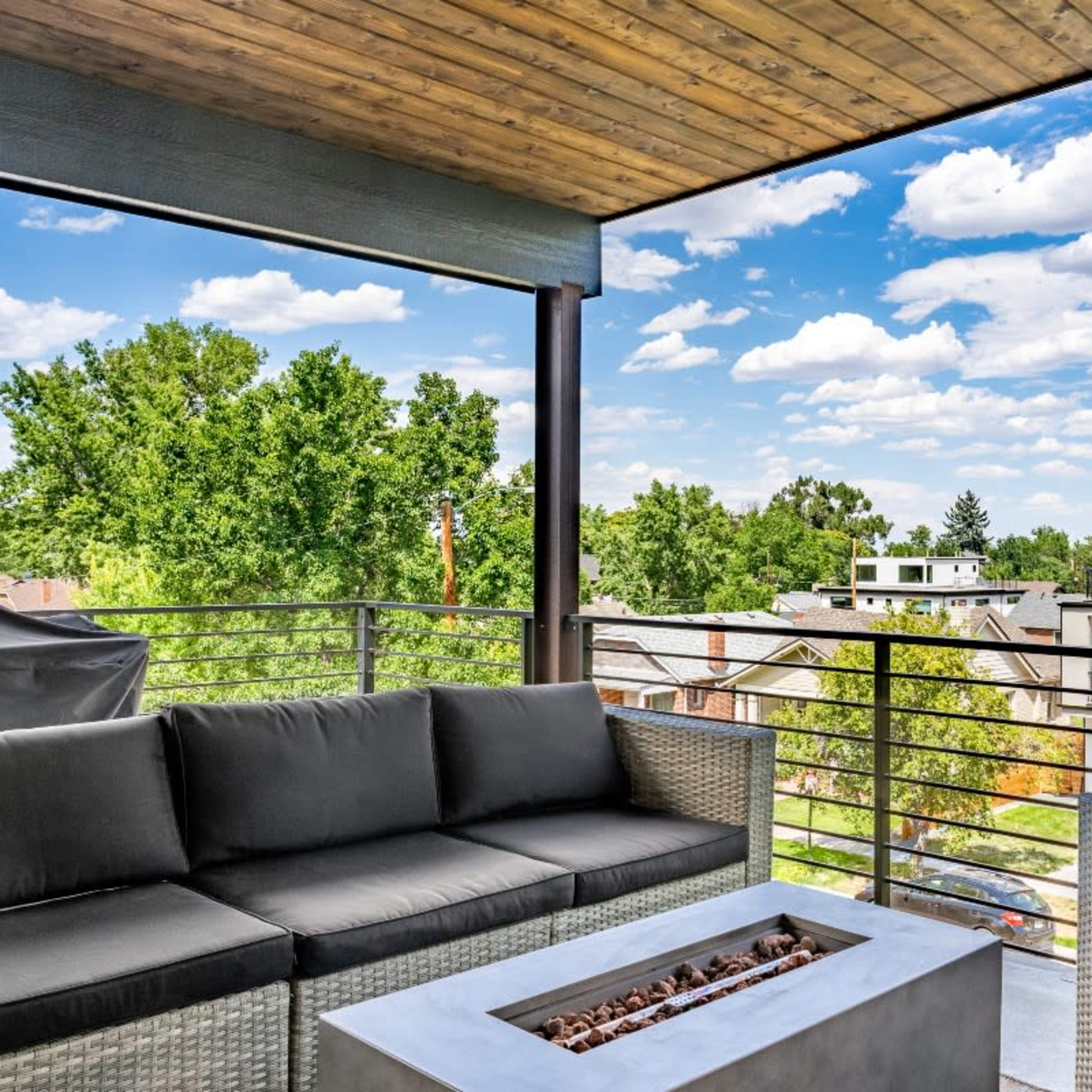 A modern outdoor seating area with a fire pit, surrounded by trees and residential buildings under a partly cloudy sky.