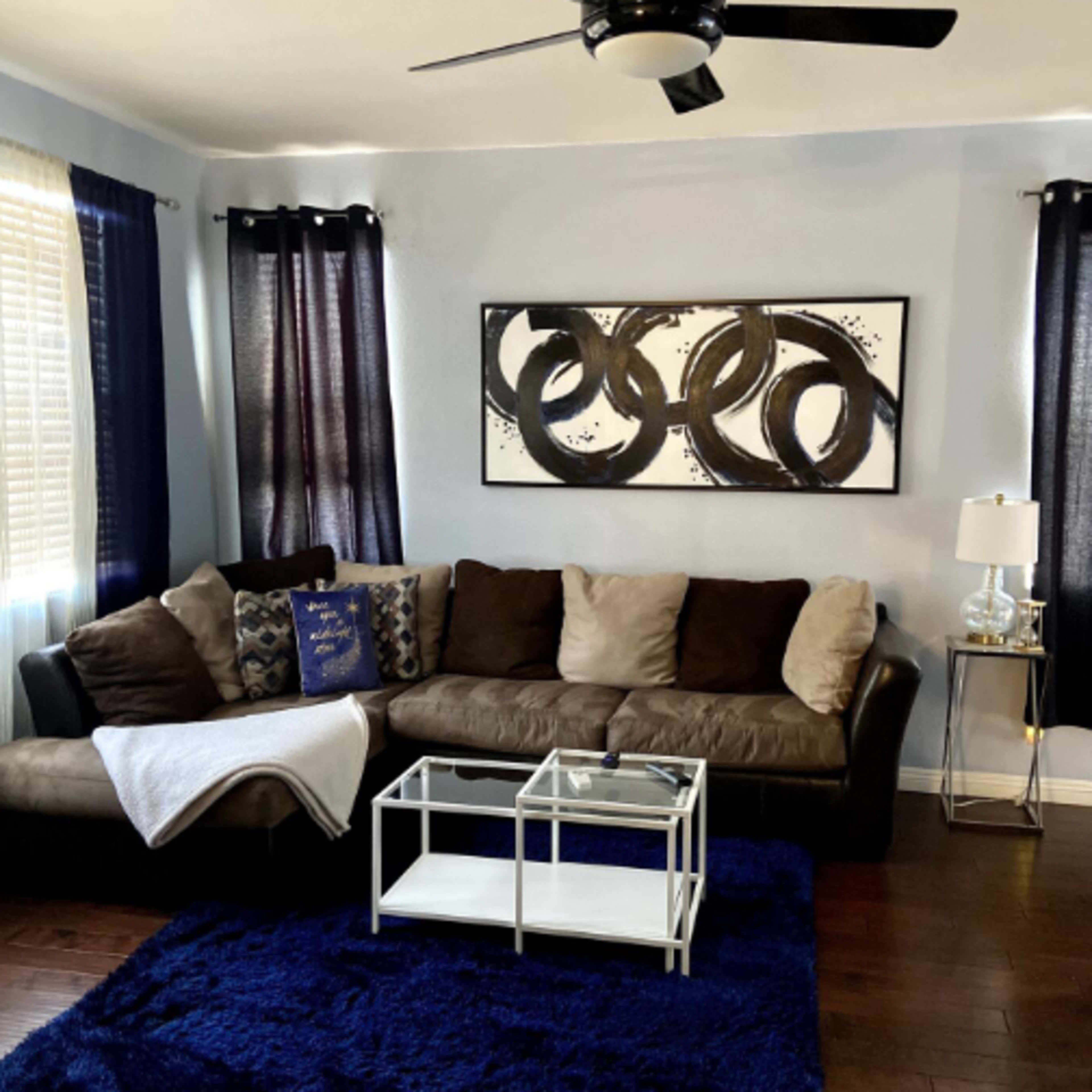 A living room features a brown sectional sofa, a glass coffee table, a blue area rug, and a wall-mounted fan, with abstract art hanging above the couch.