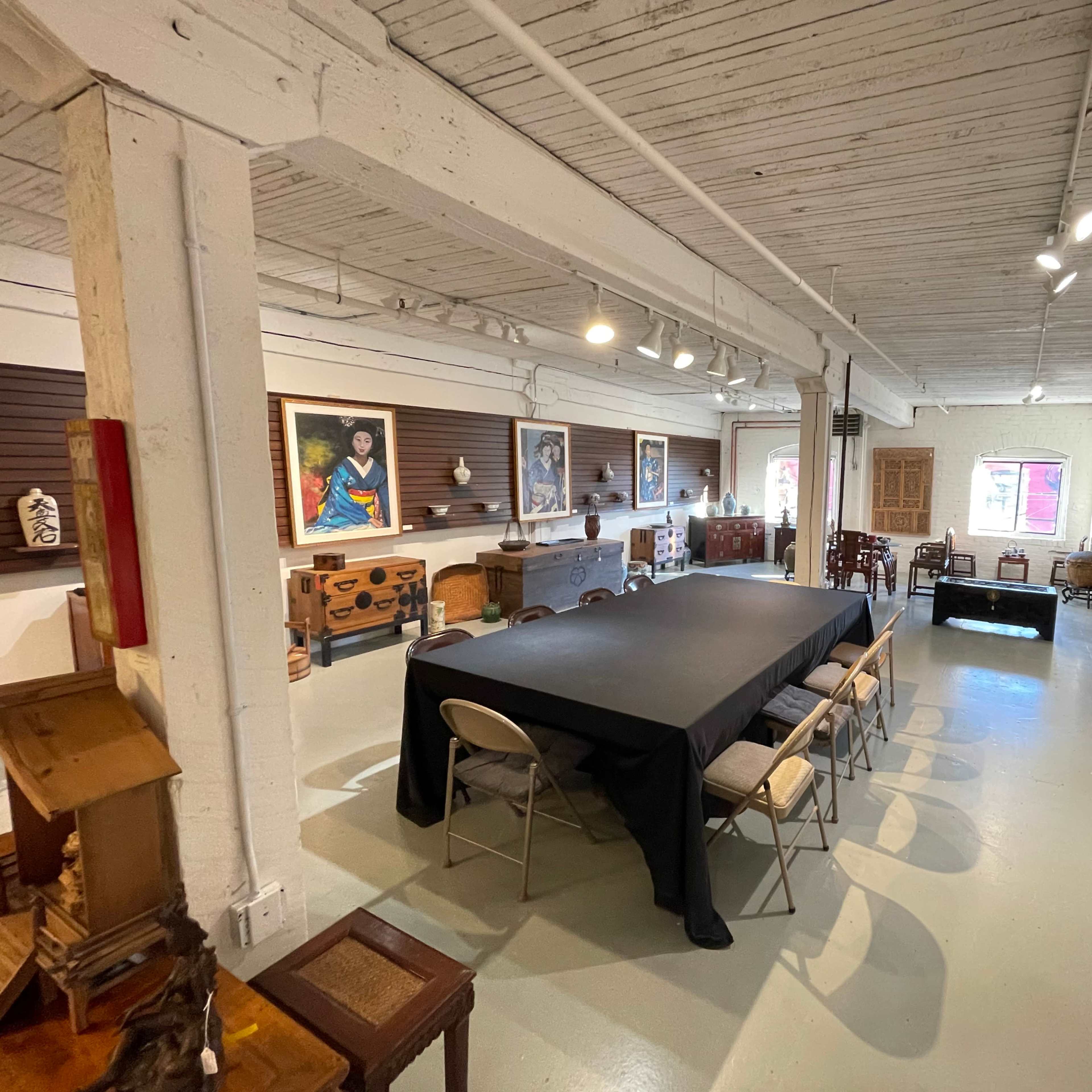 A spacious room with a large black table surrounded by chairs, artwork on the walls, and various wooden furniture pieces arranged throughout the space.