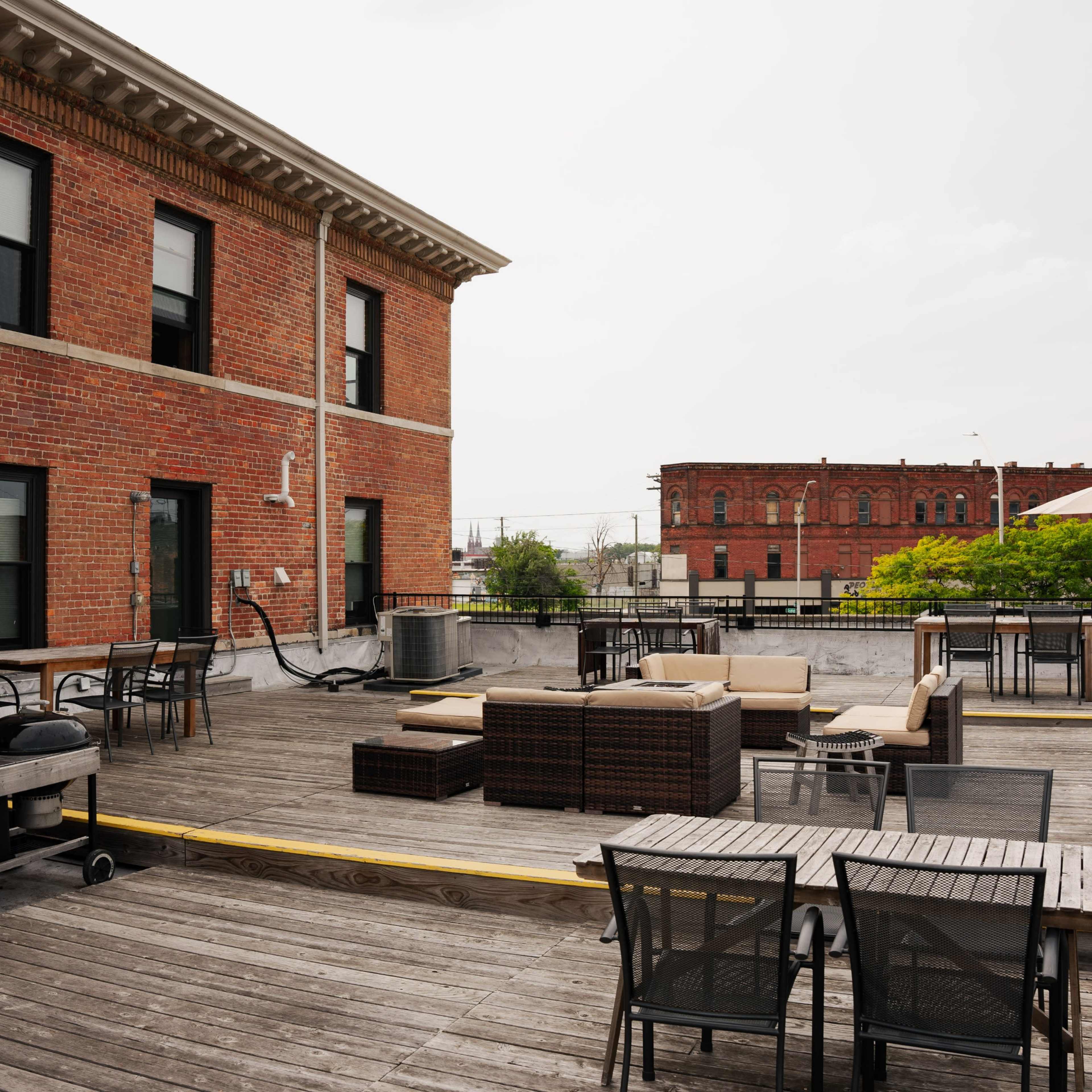 A rooftop terrace with wooden flooring, outdoor furniture, and a view of a brick building in the background.