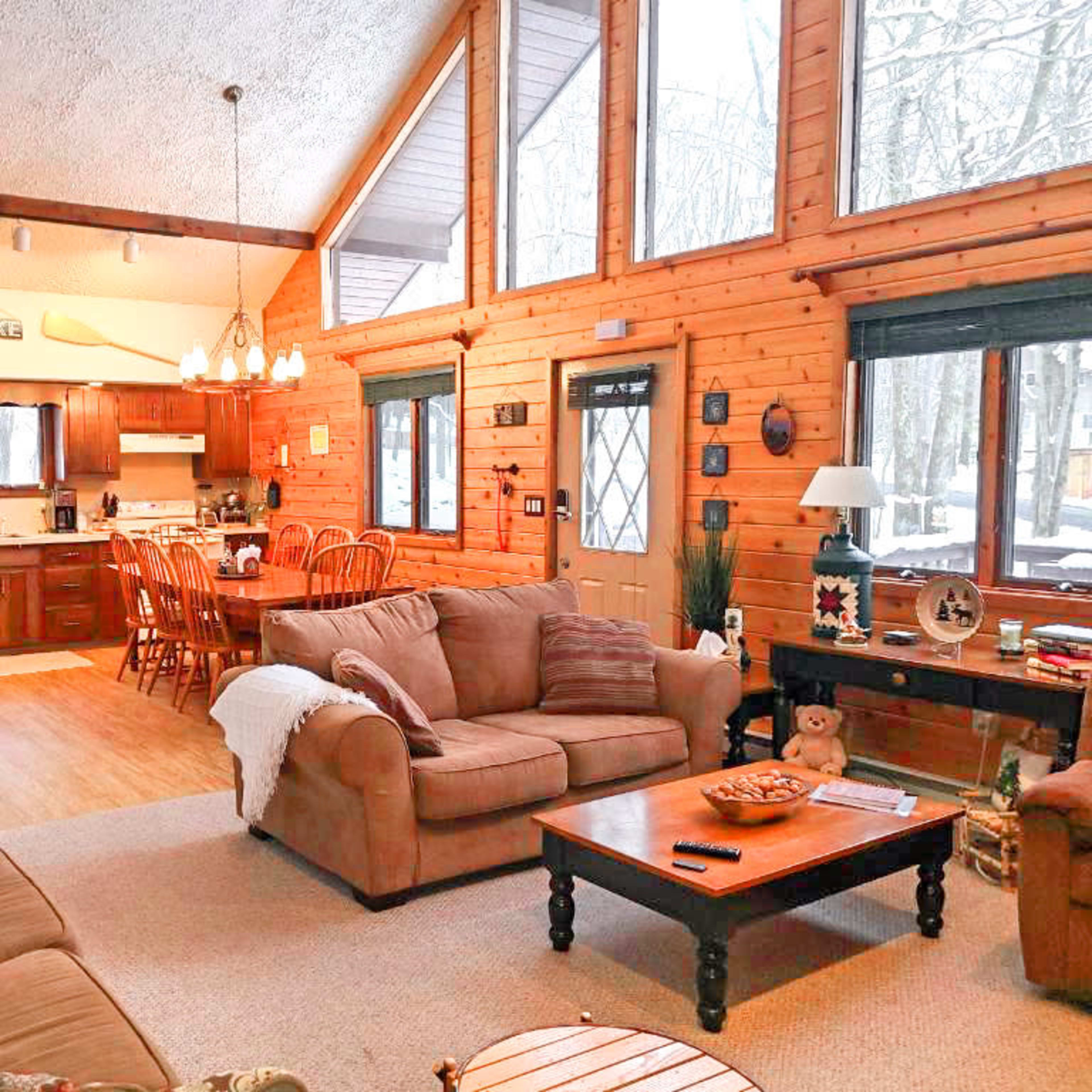 A cozy living area with wooden walls, large windows, a dining space, and a kitchen visible in the background.