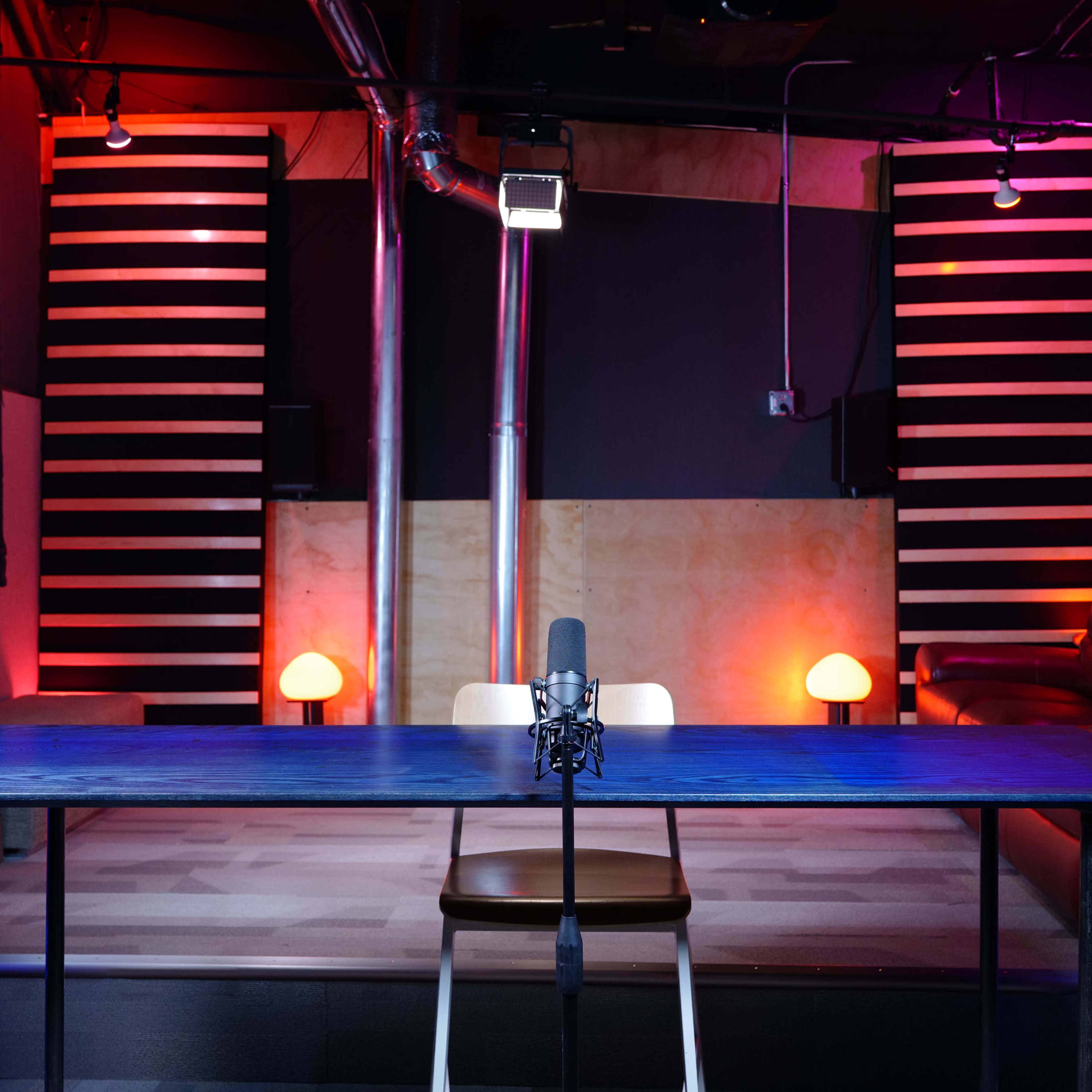 A well-lit studio with a microphone on a table facing a seating area, flanked by illuminated lamps and striped walls.