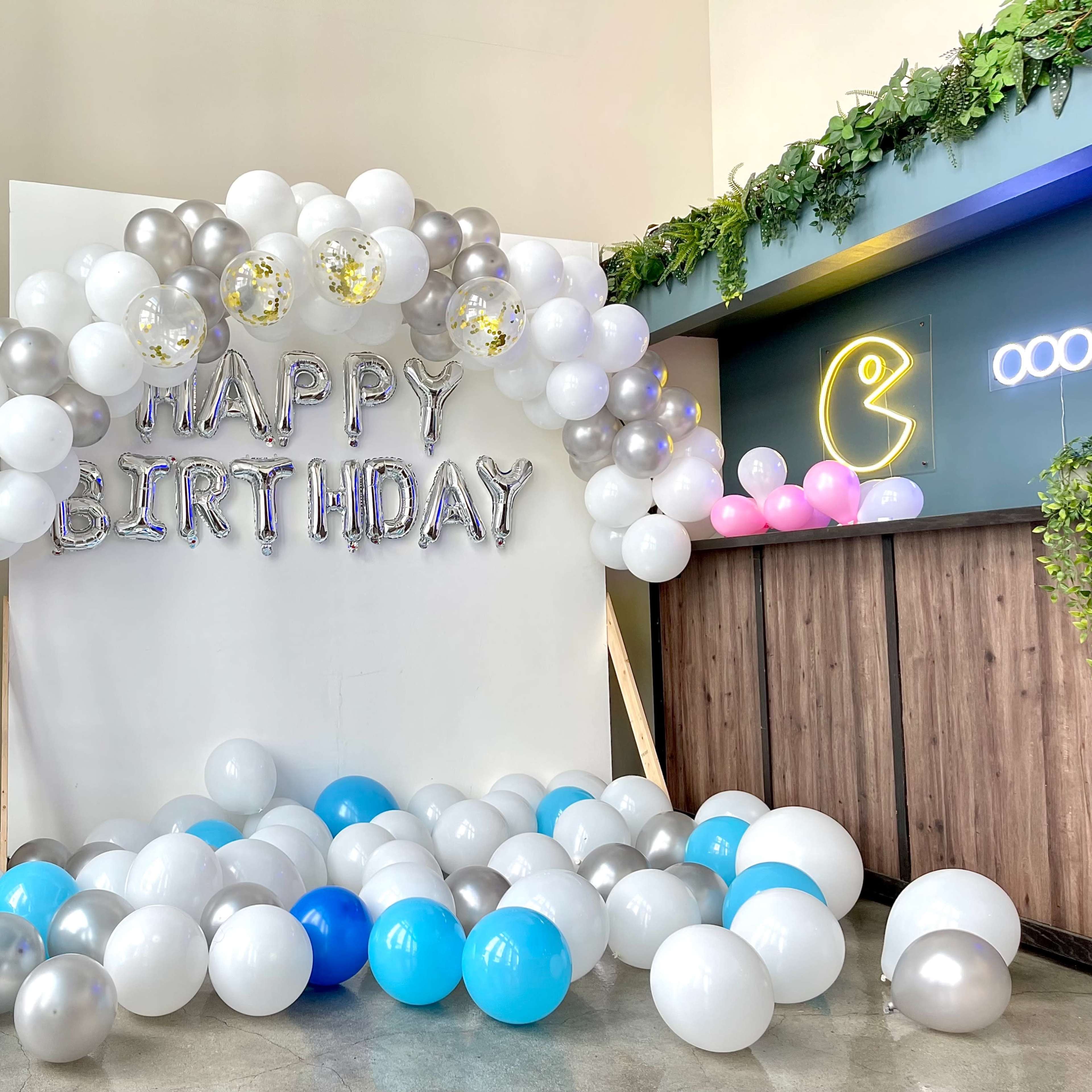 A birthday decoration setup with a "HAPPY BIRTHDAY" banner beneath a balloon arch, surrounded by a mix of blue, silver, and white balloons on the floor.