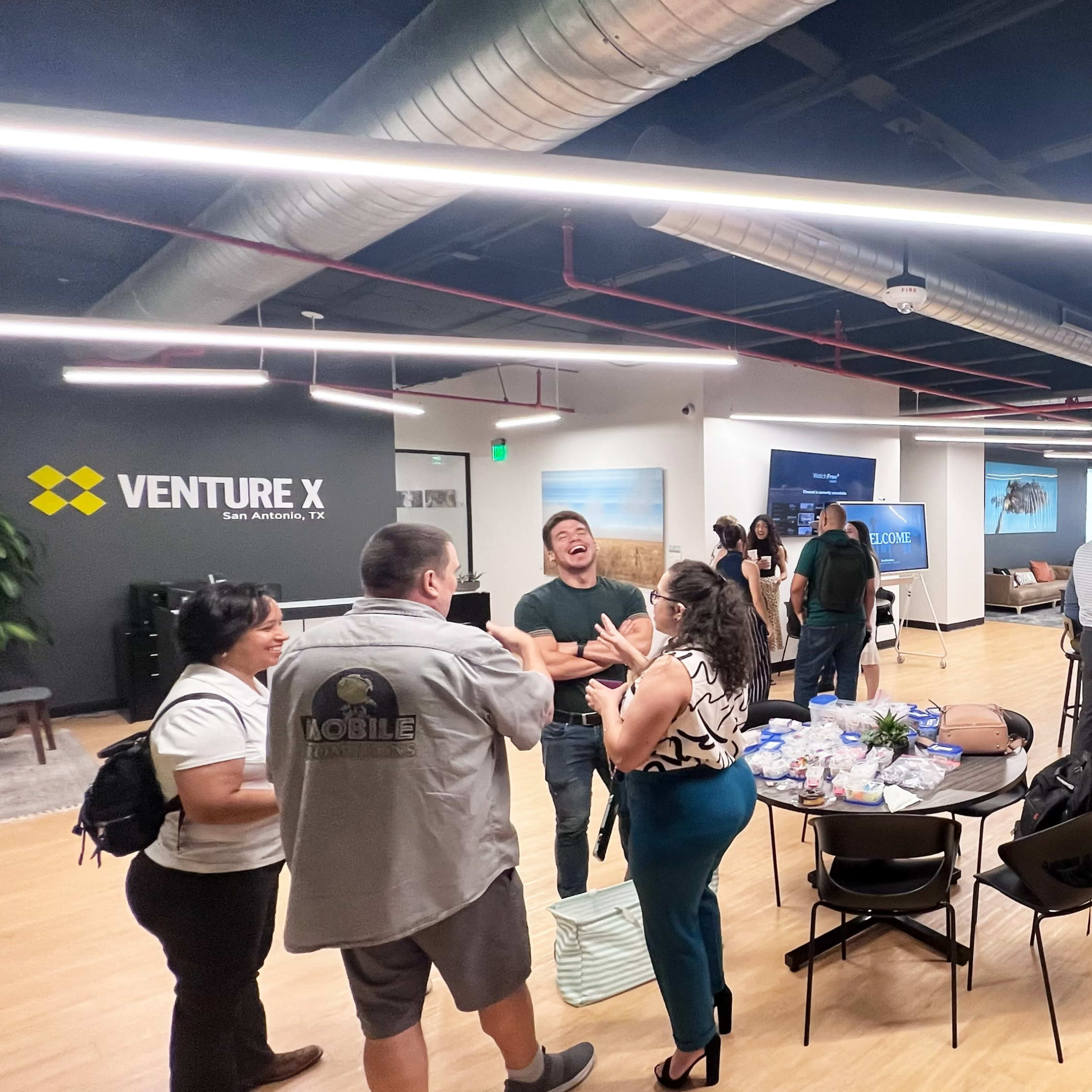 A group of people are networking at a business event in a modern office space with seating areas and a refreshment table.