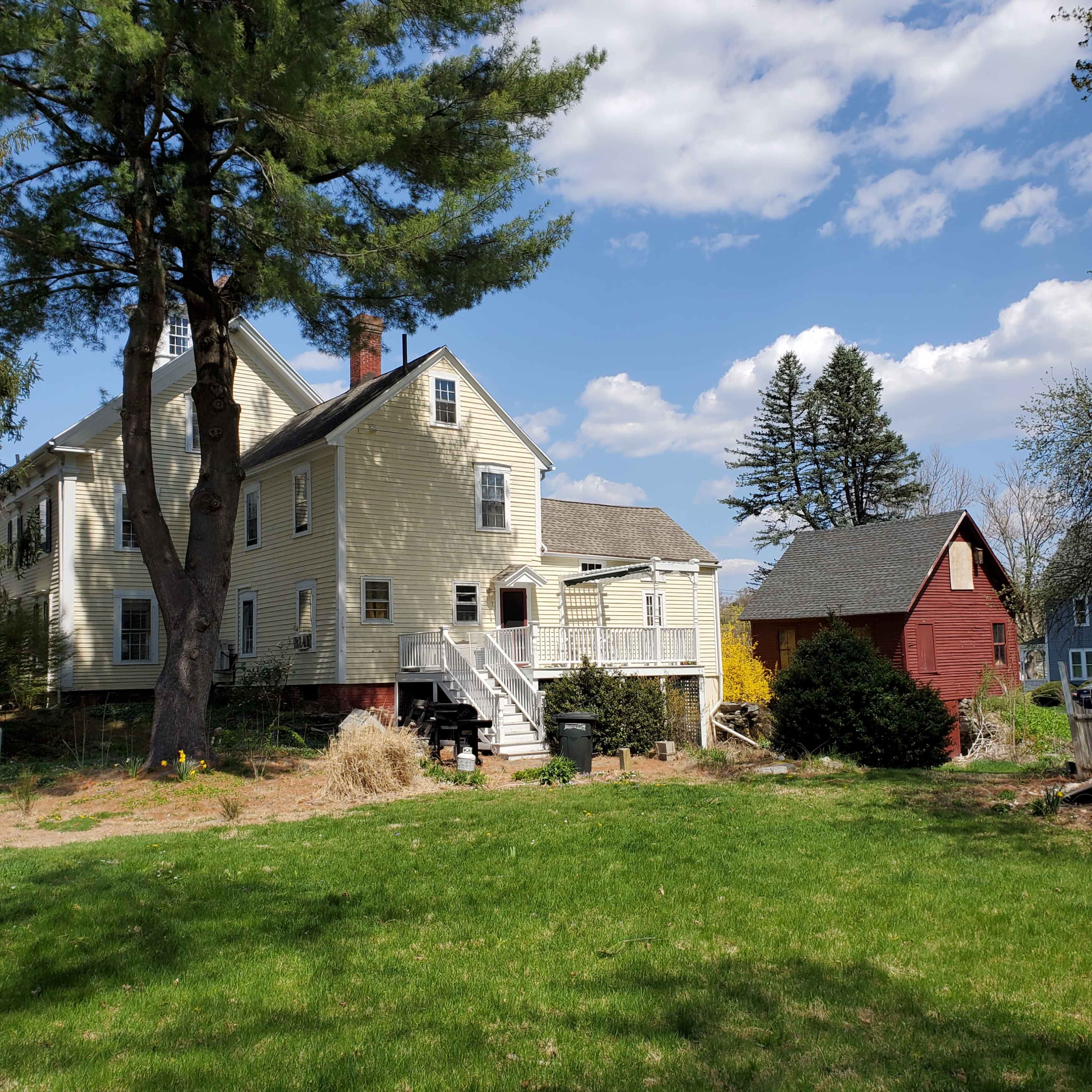 A yellow two-story house with a white porch is situated in a grassy yard, alongside a red shed and surrounded by trees under a partly cloudy sky.