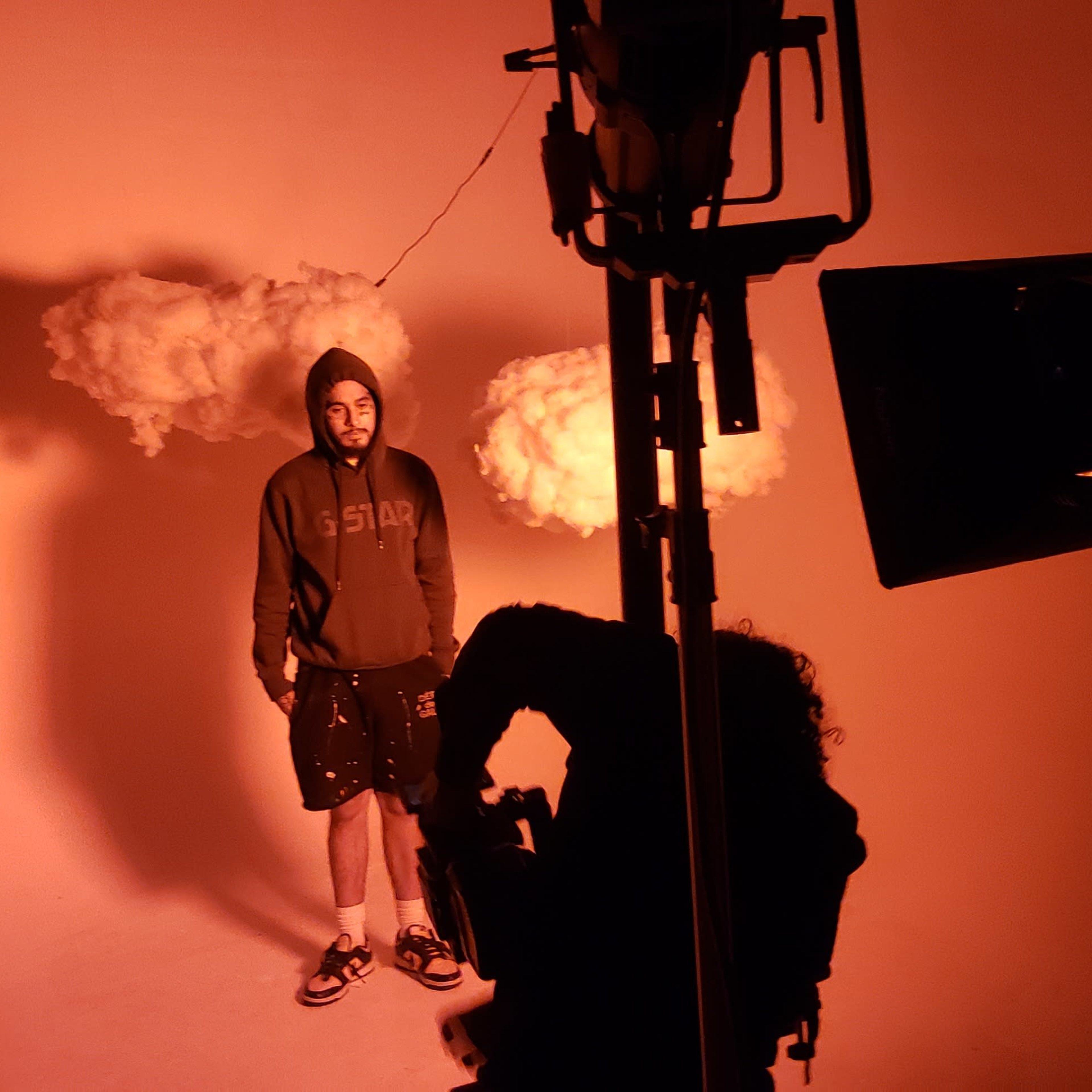 A person wearing a hoodie stands under a suspended cloud prop, while two individuals are positioned nearby with a camera and lighting equipment.
