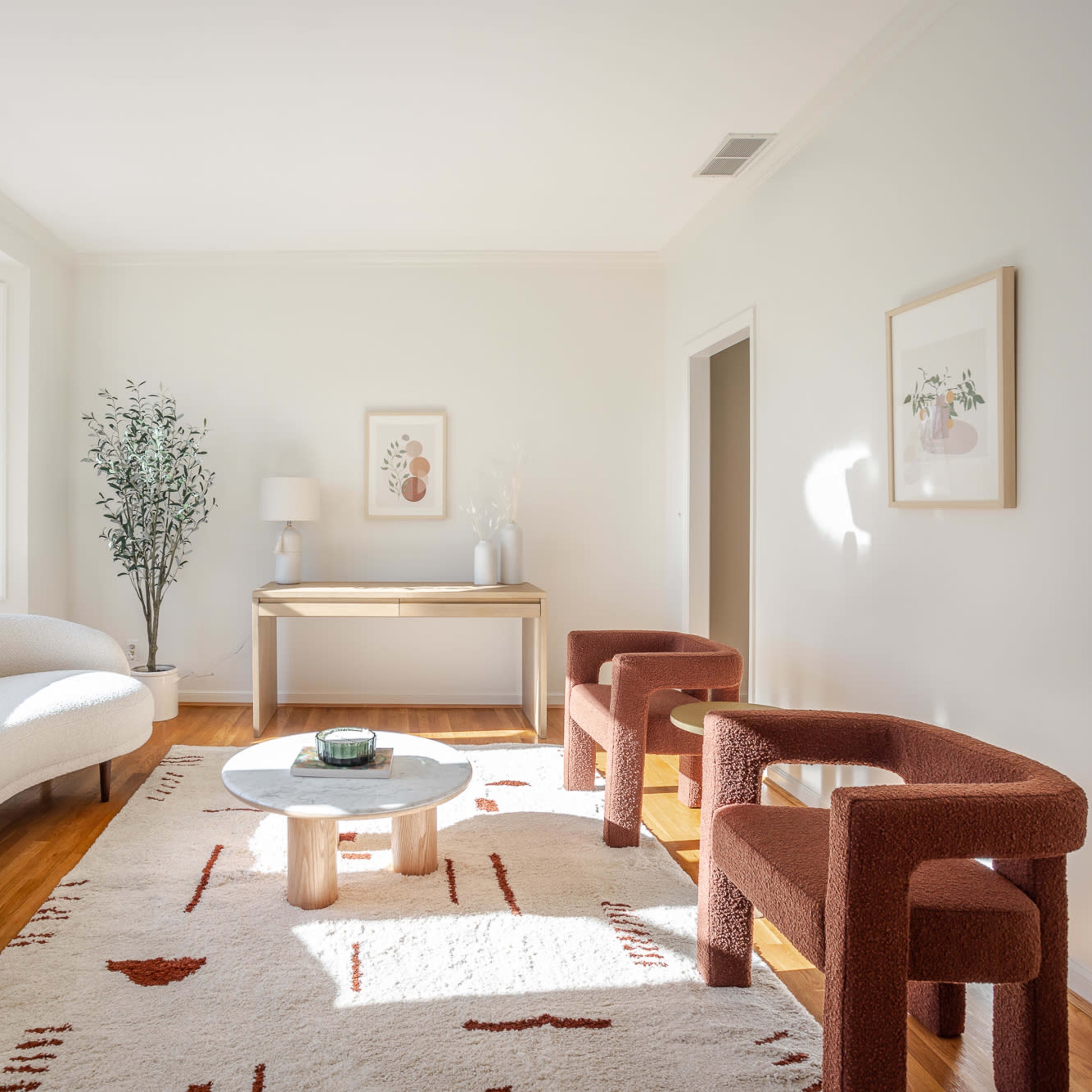 A bright, minimalist living room features a curved white sofa, two red upholstered chairs, a round coffee table, and a console table against a light-colored wall.