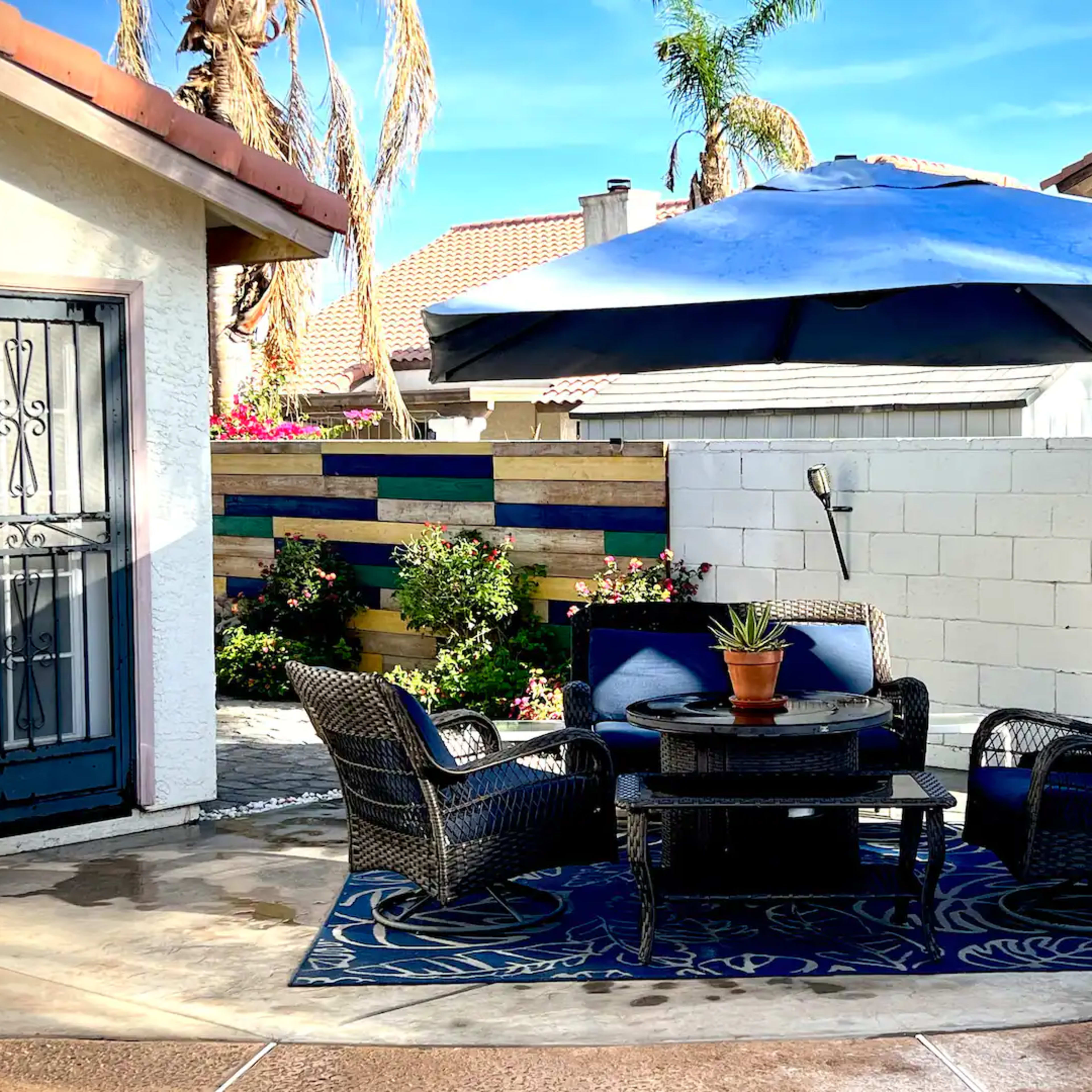 A shaded patio area features a round table surrounded by four chairs, with a potted plant on the table and colorful landscaping in the background.