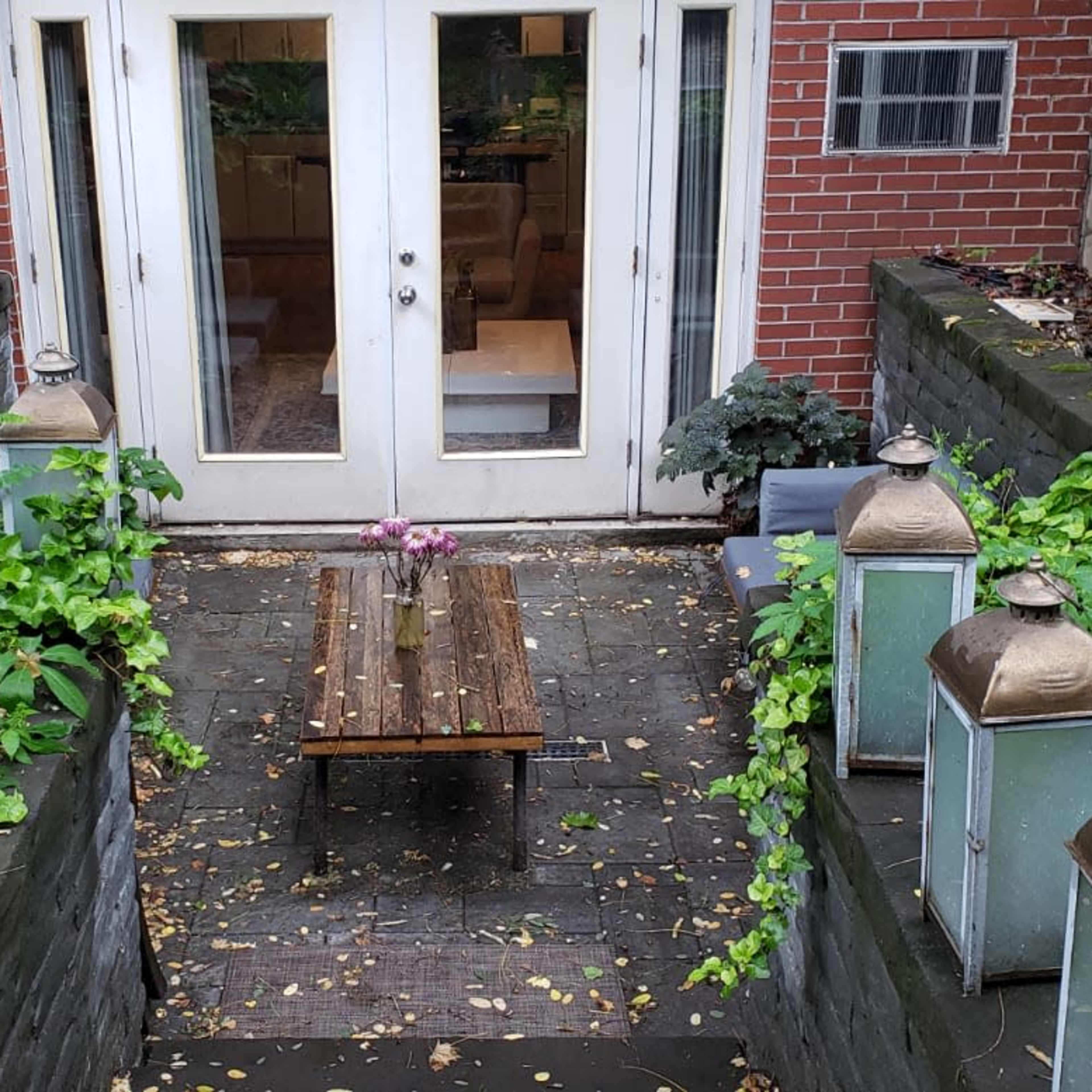 A small, garden patio with a narrow wooden bench surrounded by greenery, leading to a set of double doors.