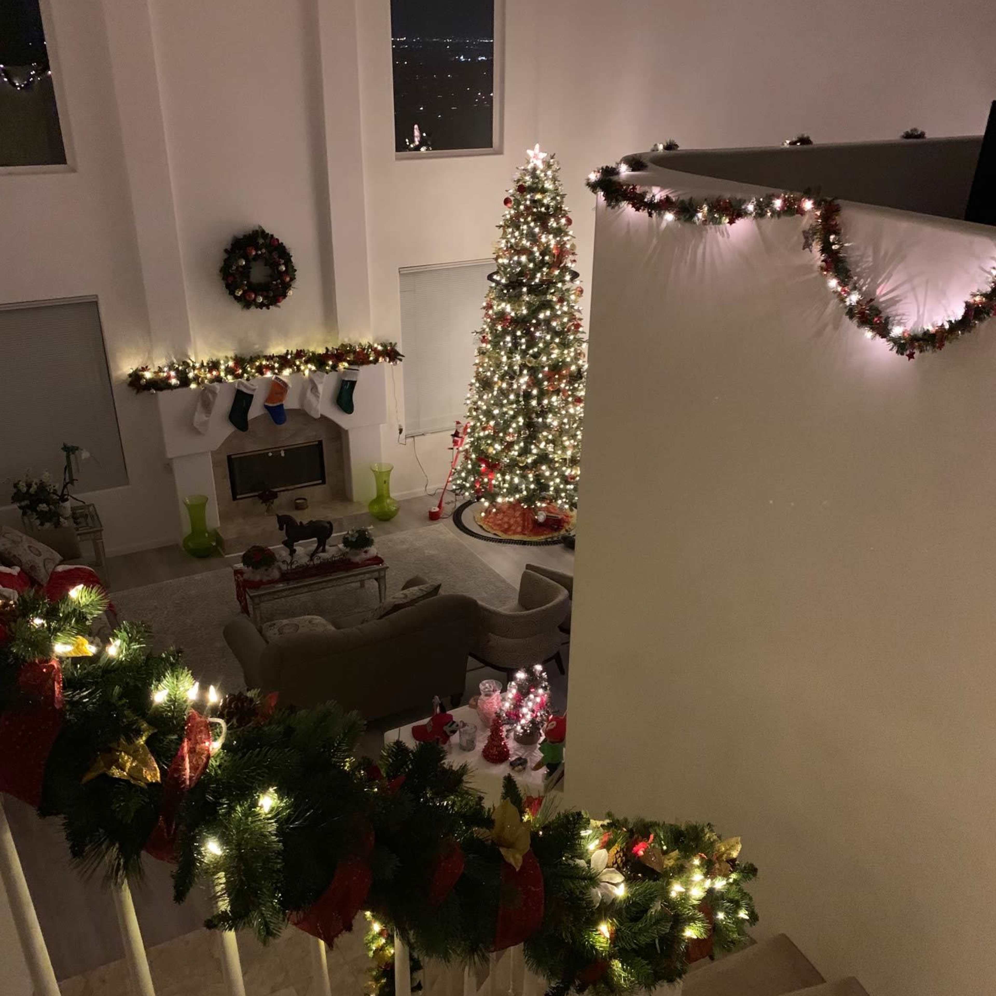 A decorated living room with a Christmas tree, stockings on the fireplace, and garlands lighting up a staircase.