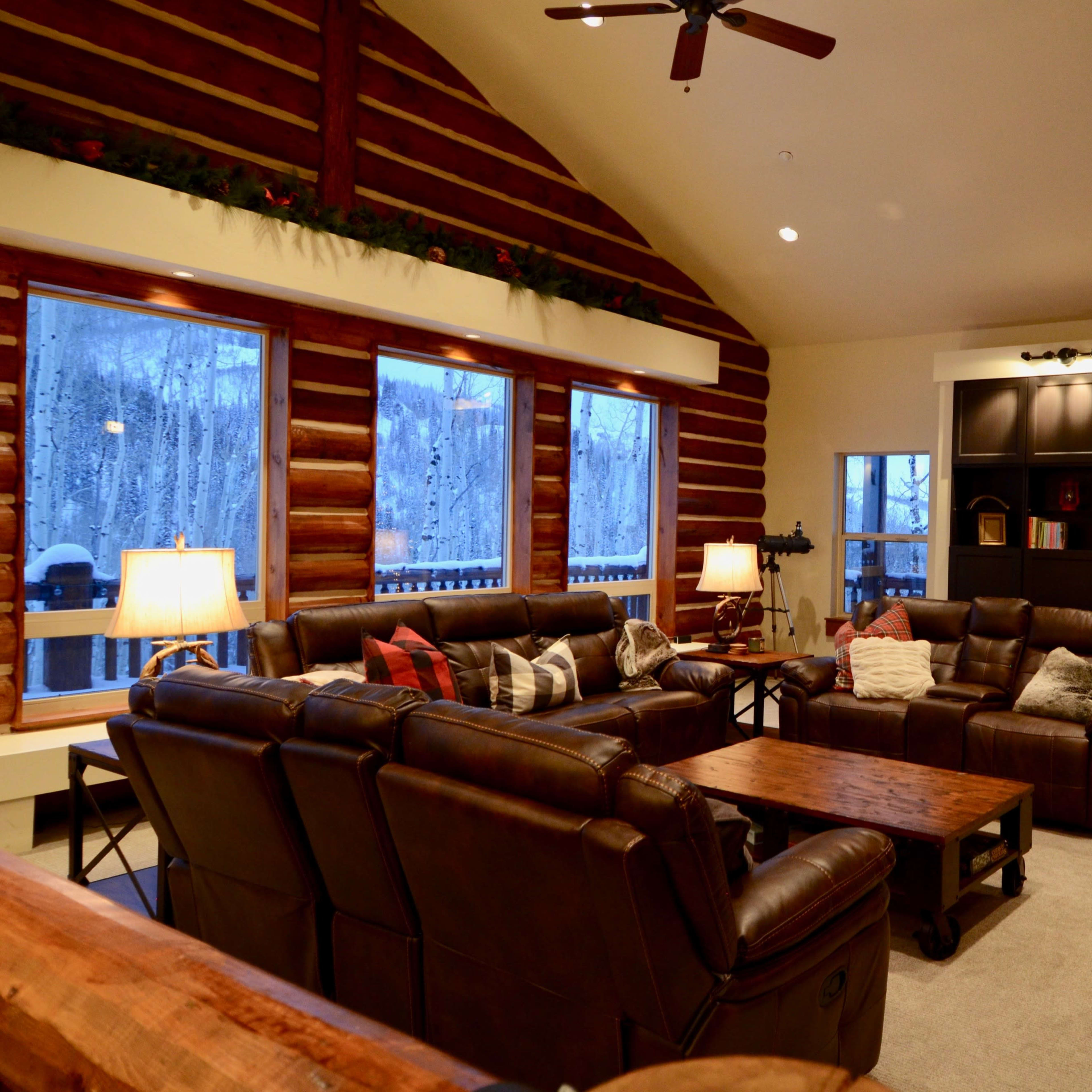 The cozy living room features leather sofas, a wooden coffee table, large windows showcasing a snowy landscape, and a decorated Christmas tree.