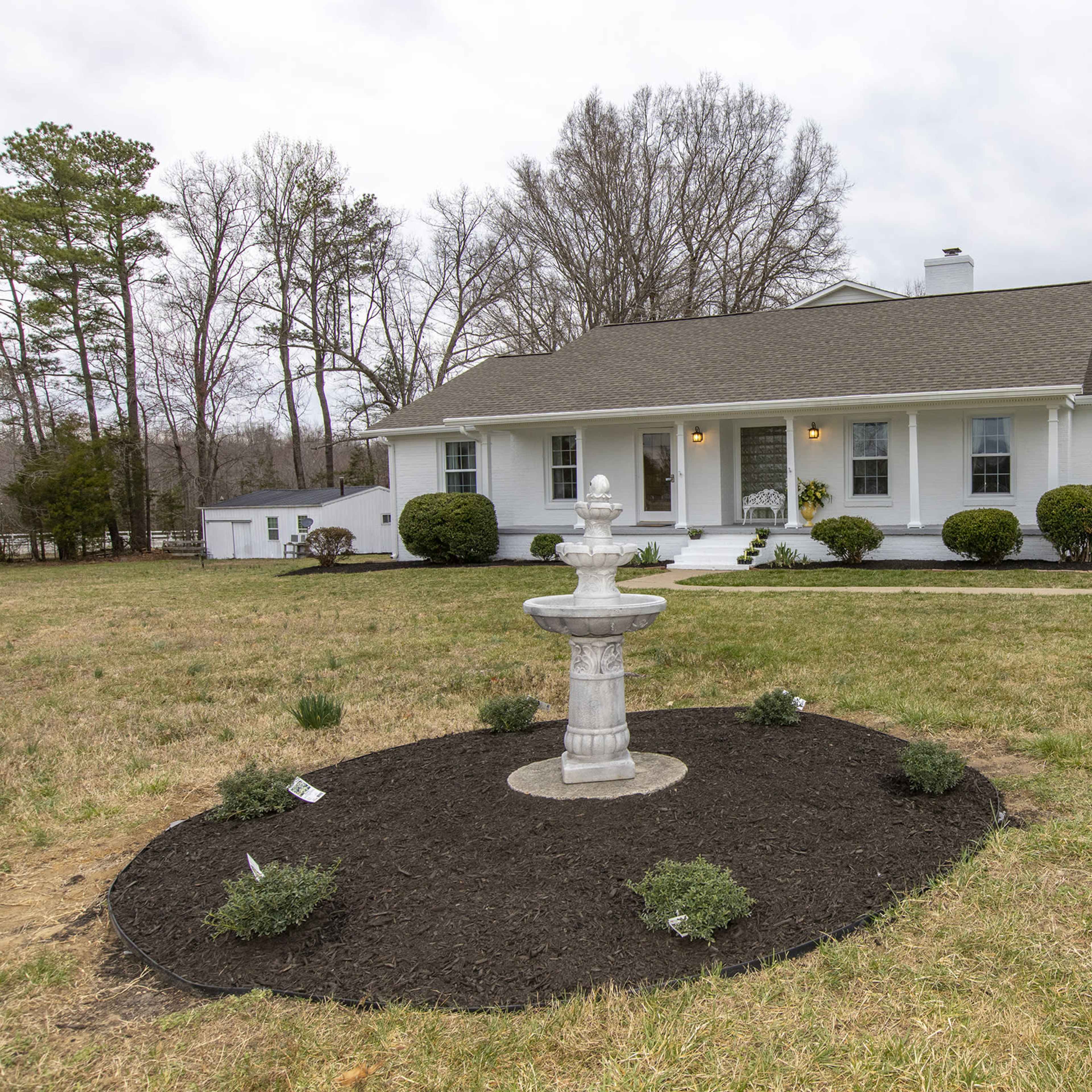 A white, single-story house with a gray roof is surrounded by landscaped grass and shrubs, featuring a stone fountain in the foreground.
