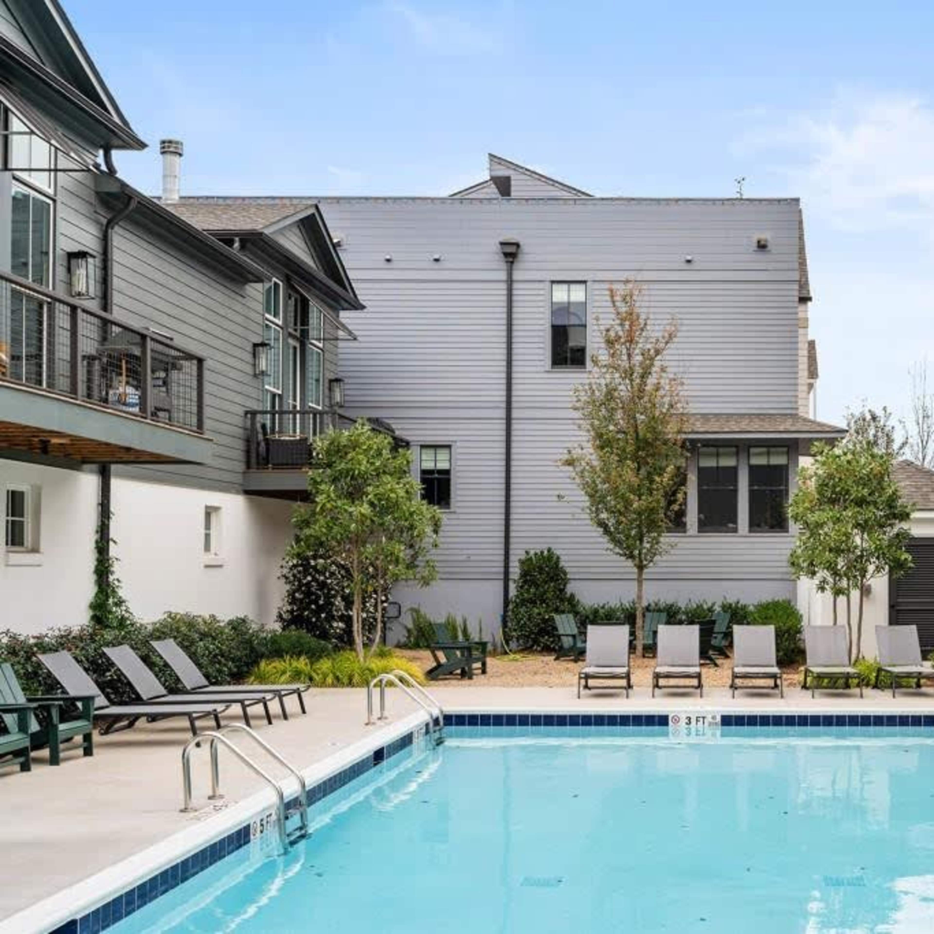 A swimming pool is surrounded by chaise lounges and small trees, with two multi-story buildings in the background.