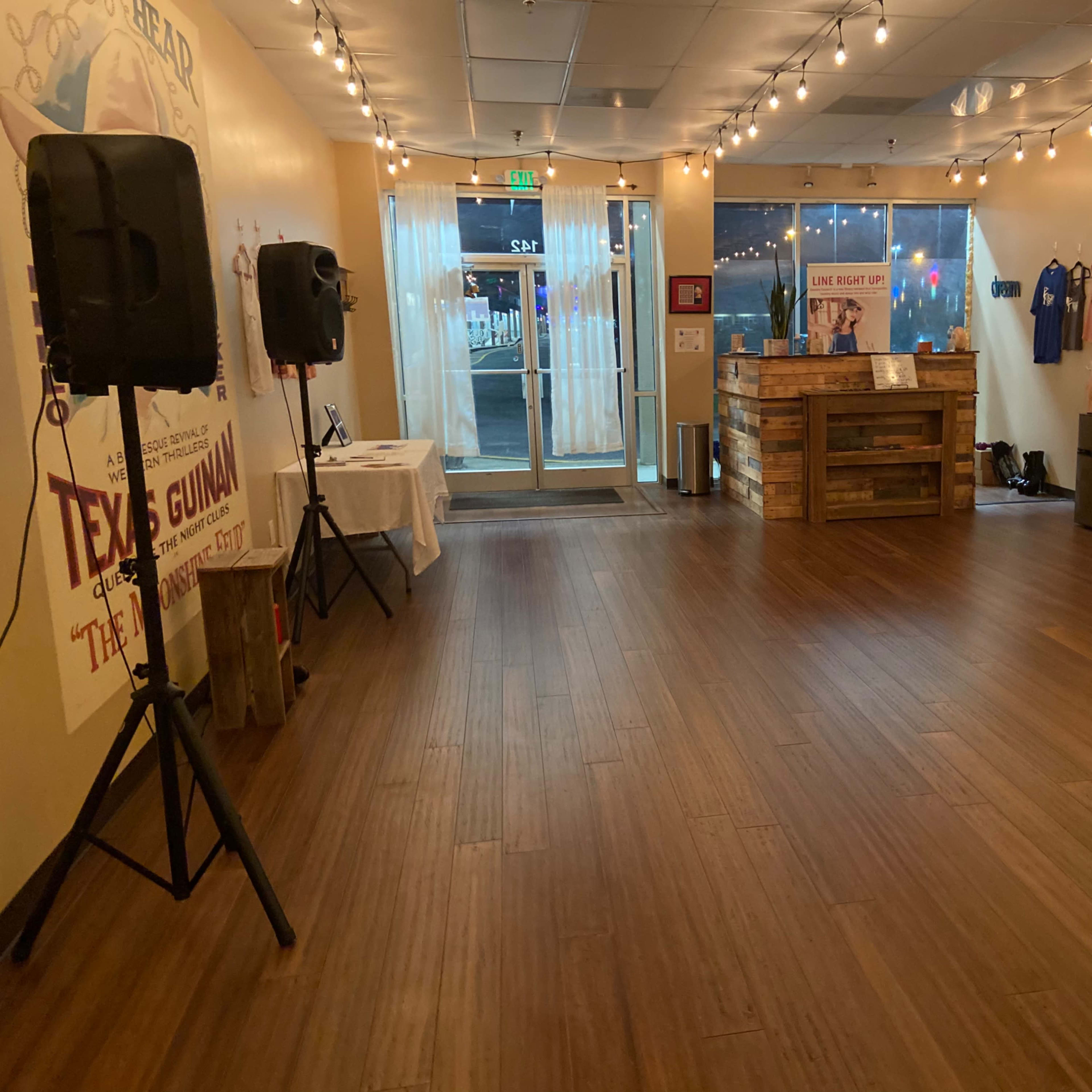 An empty dance studio with wooden flooring, speakers on stands, and a reception area featuring a wooden counter and mirrored walls.