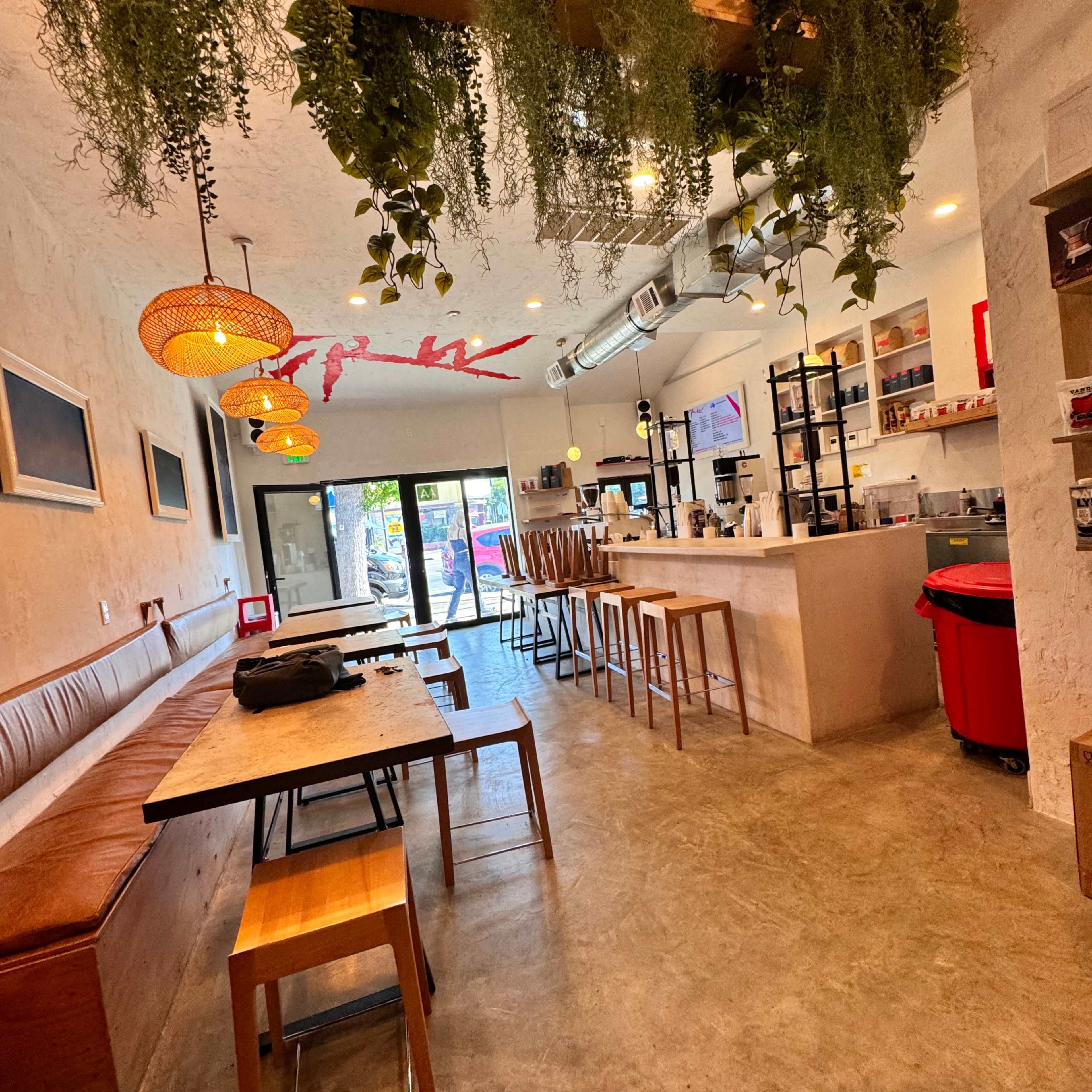 A modern café interior with a wooden ceiling structure adorned with hanging plants, wooden tables, and a counter with seating arrangements.