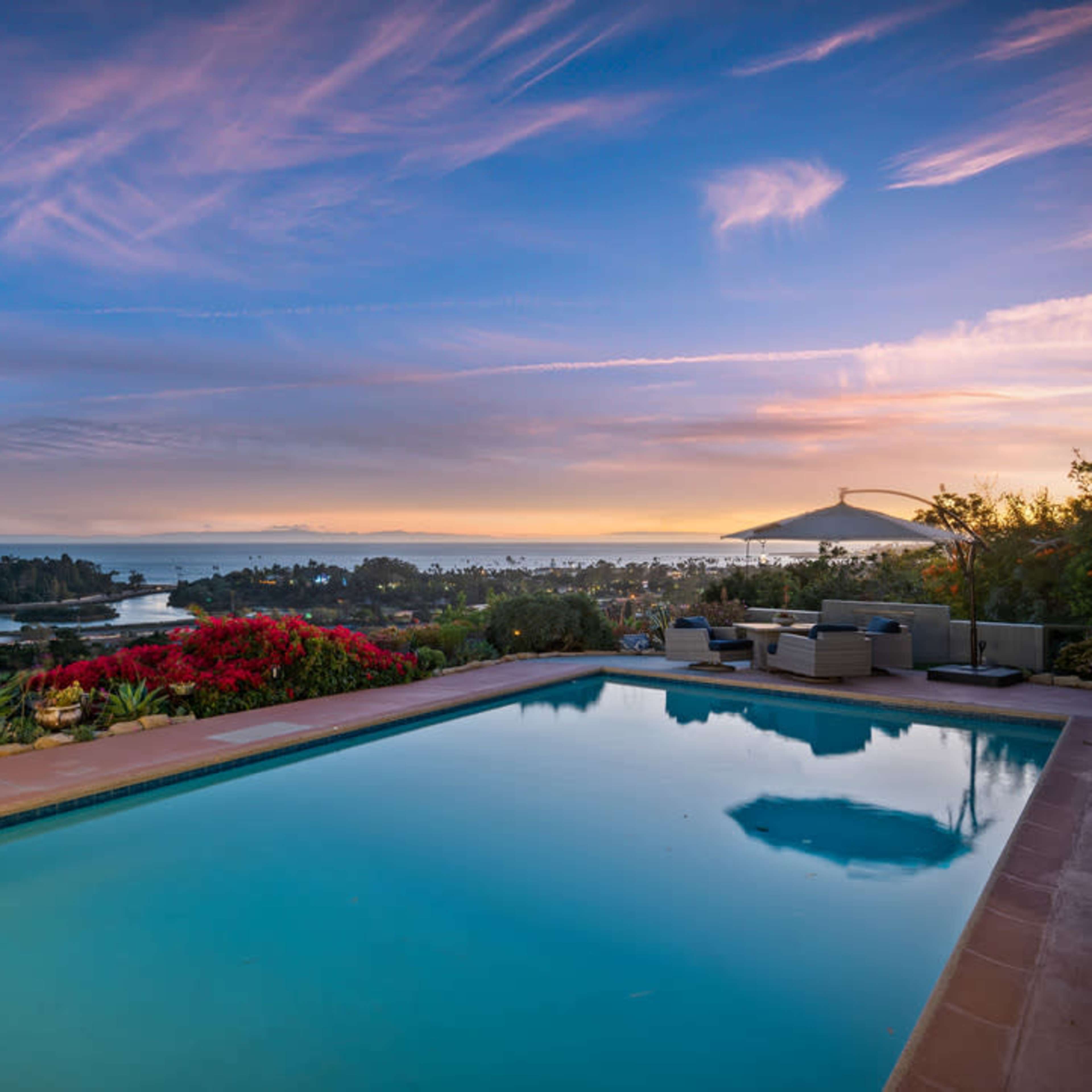 A swimming pool reflects the colorful sunset over a coastal landscape with lush greenery and flowering plants.