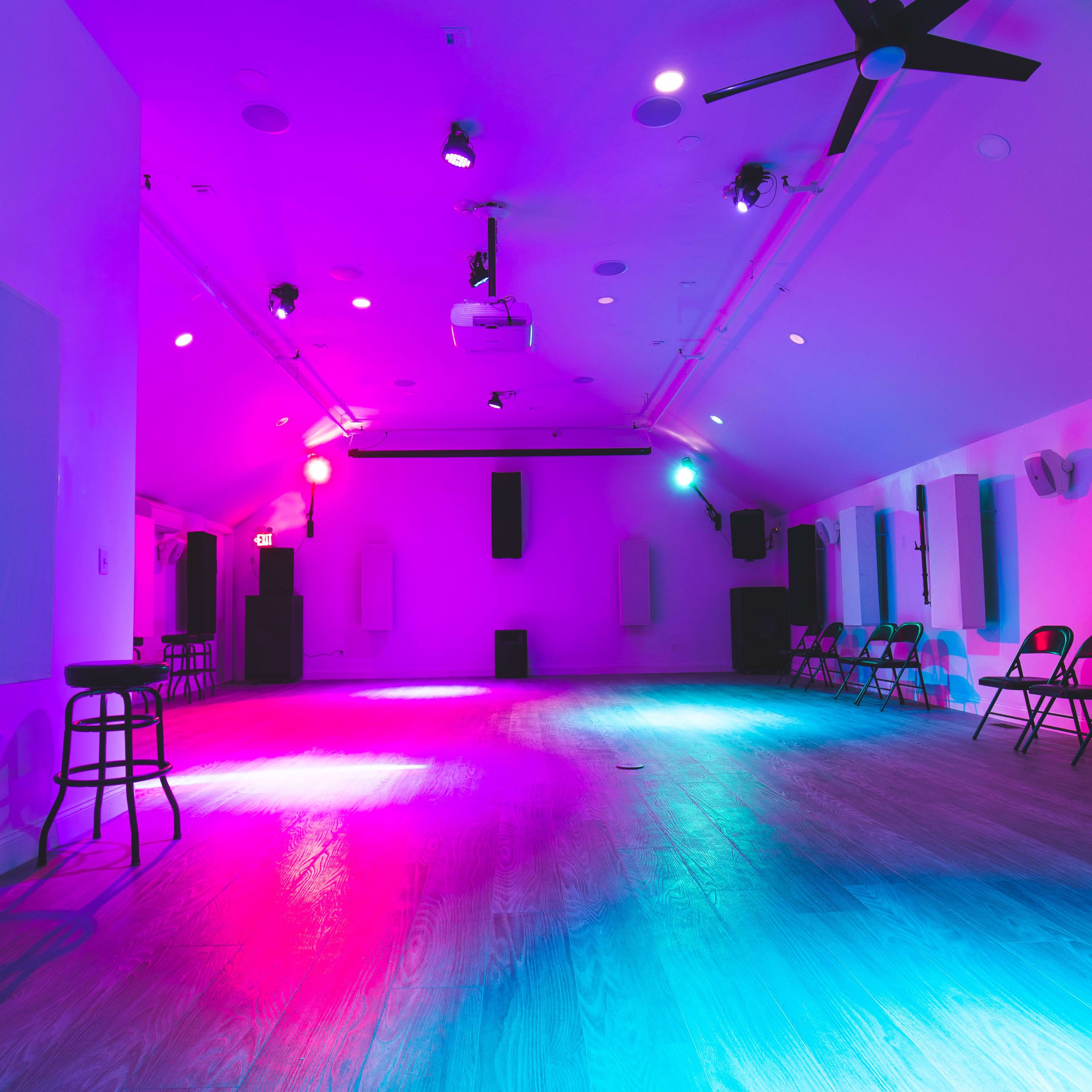 An empty room with wooden floors, illuminated by purple and blue lights, featuring folding chairs along the edges.
