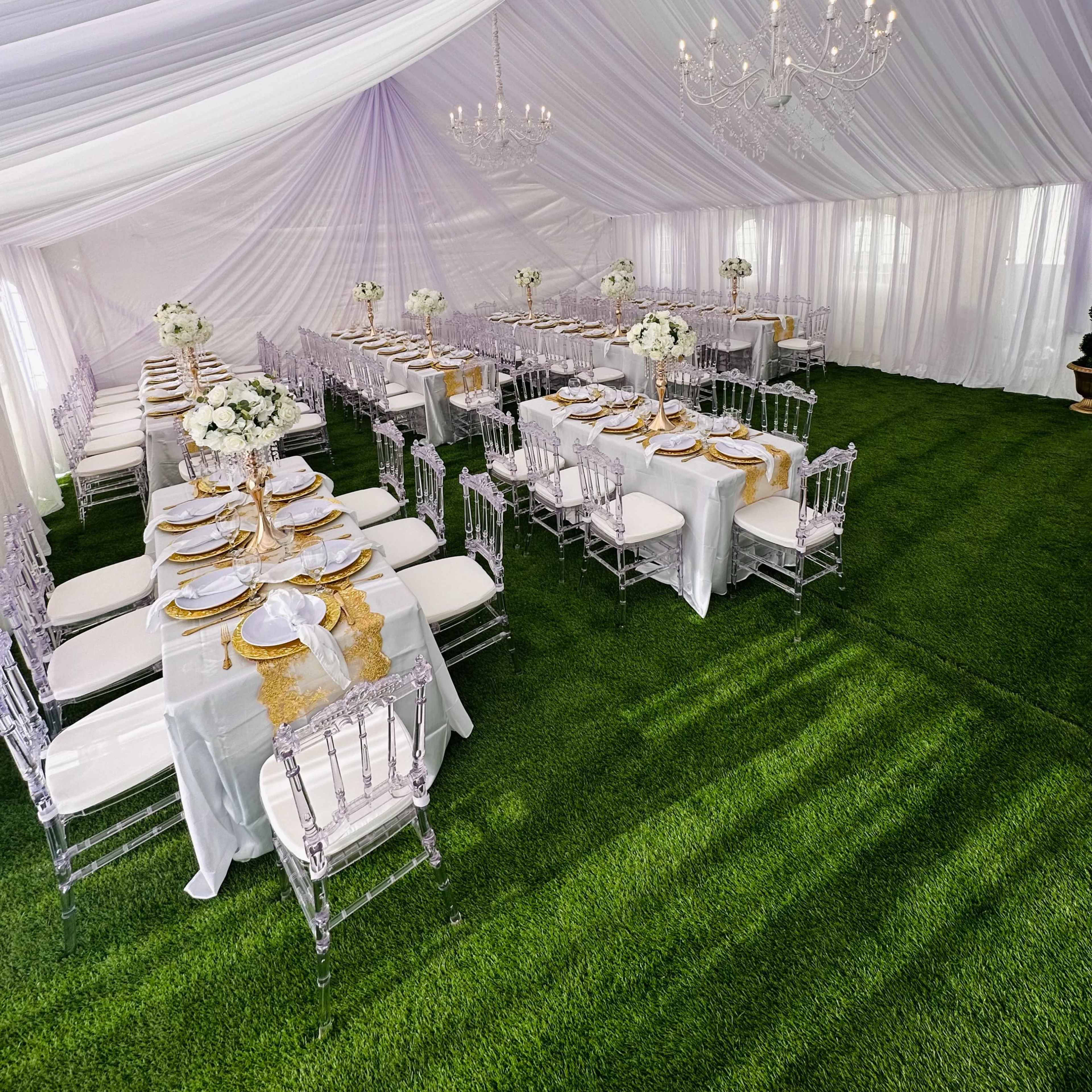A large tented space features elegantly set dining tables with white tablecloths, gold accents, and floral centerpieces, surrounded by artificial grass.