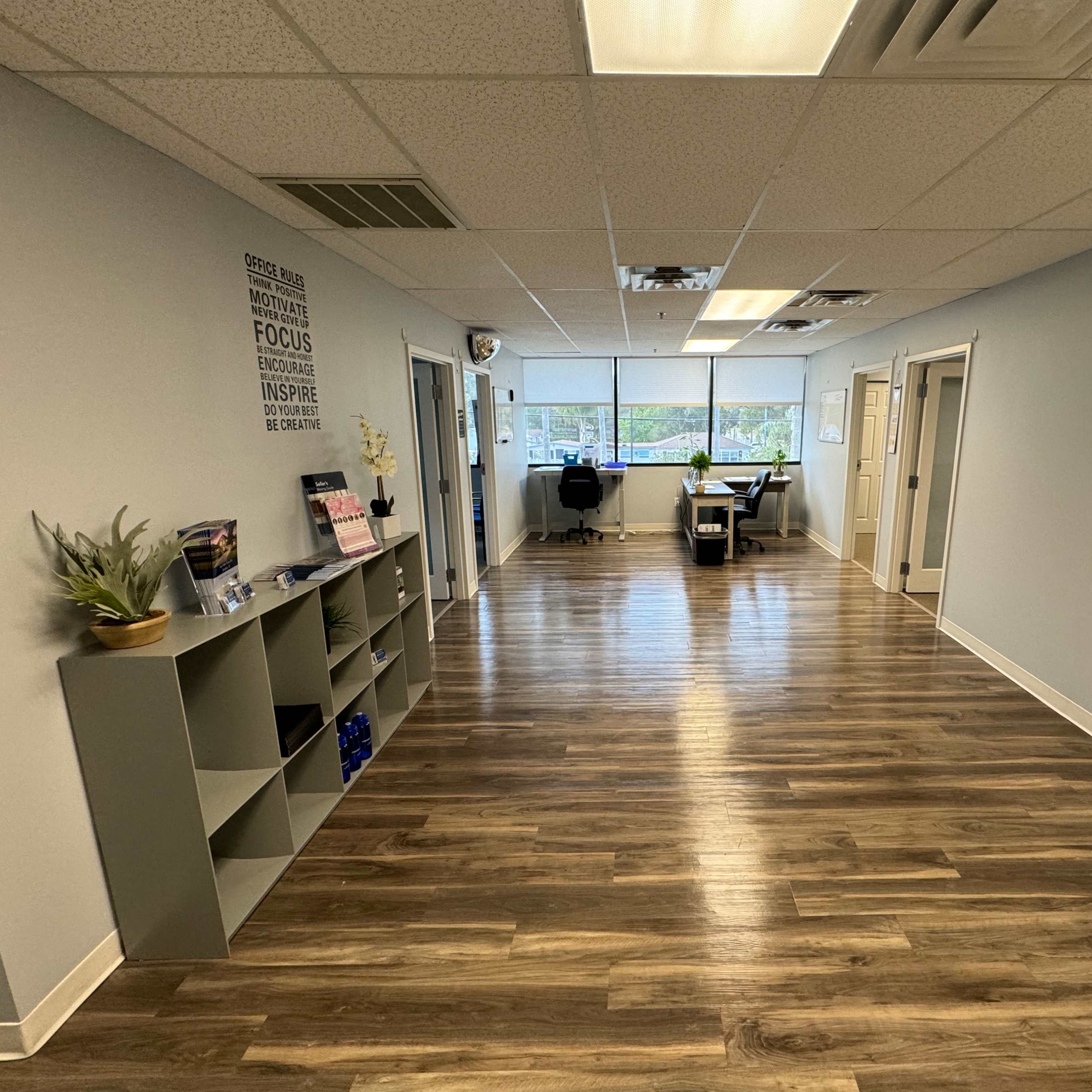 A well-lit hallway with grey walls, wooden flooring, and office doors leading to a workspace at the end.
