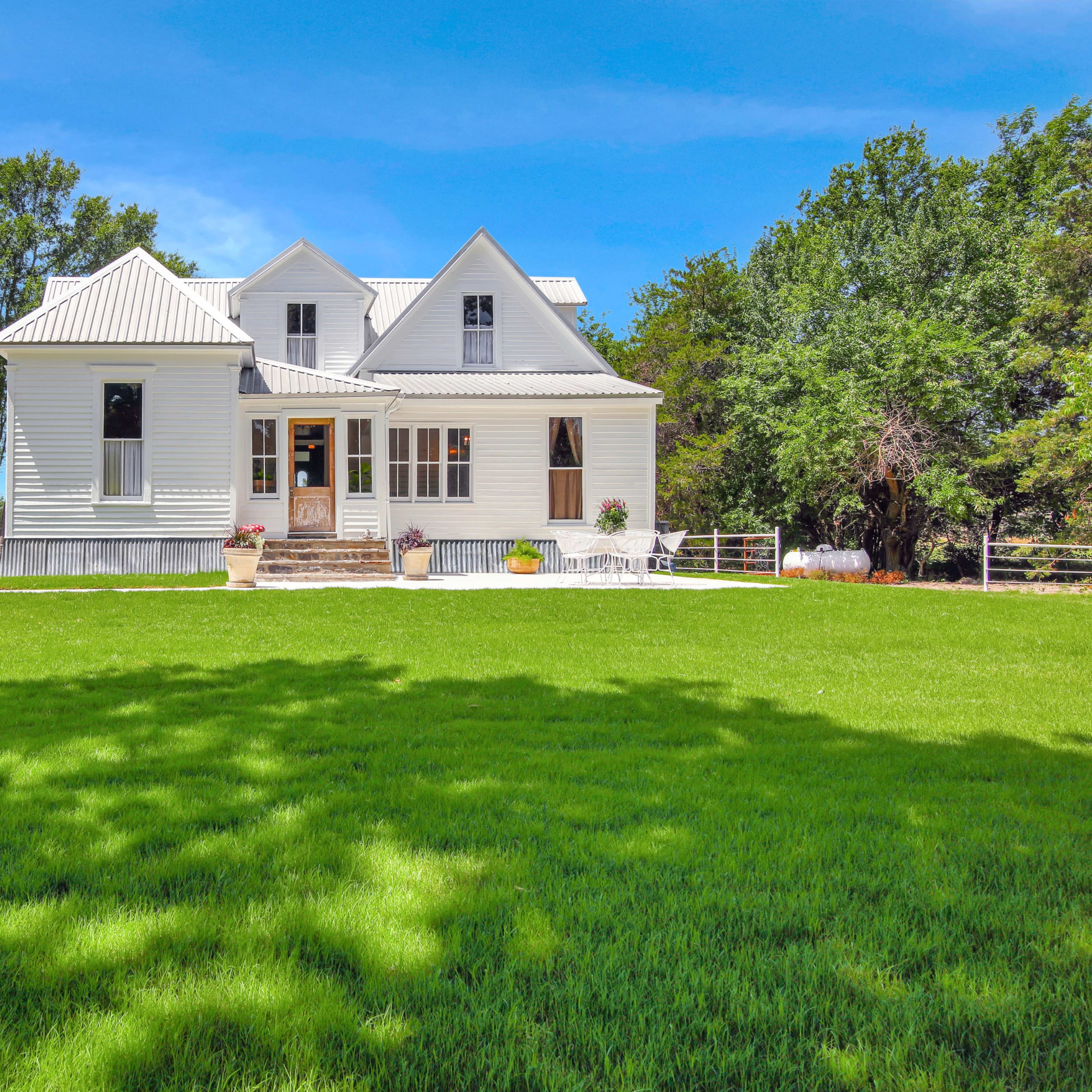 A white farmhouse with a metal roof sits on a well-kept lawn surrounded by trees.
