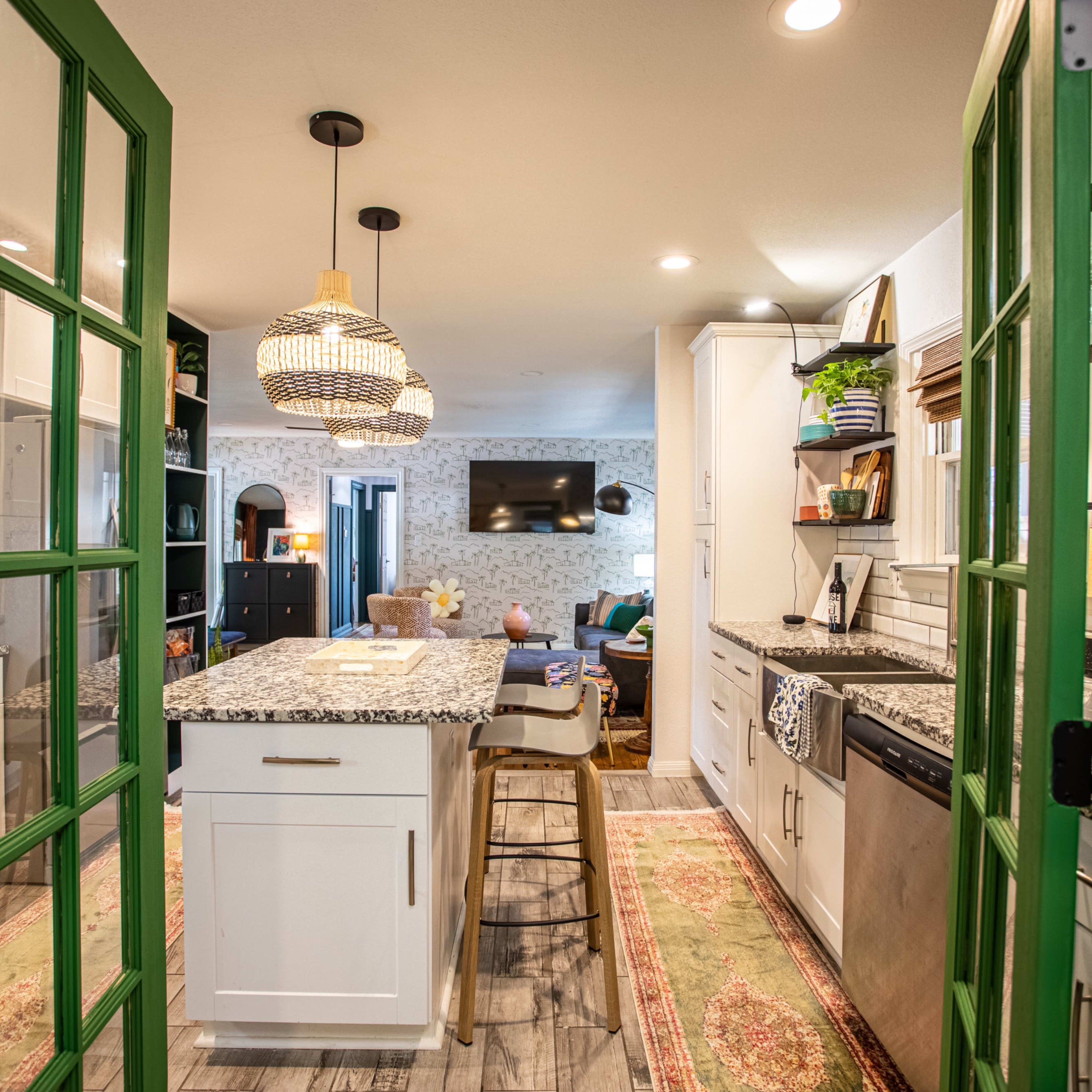 A modern kitchen viewed through green-framed double doors, featuring white cabinetry, a central island with bar seating, and a pendant light above.