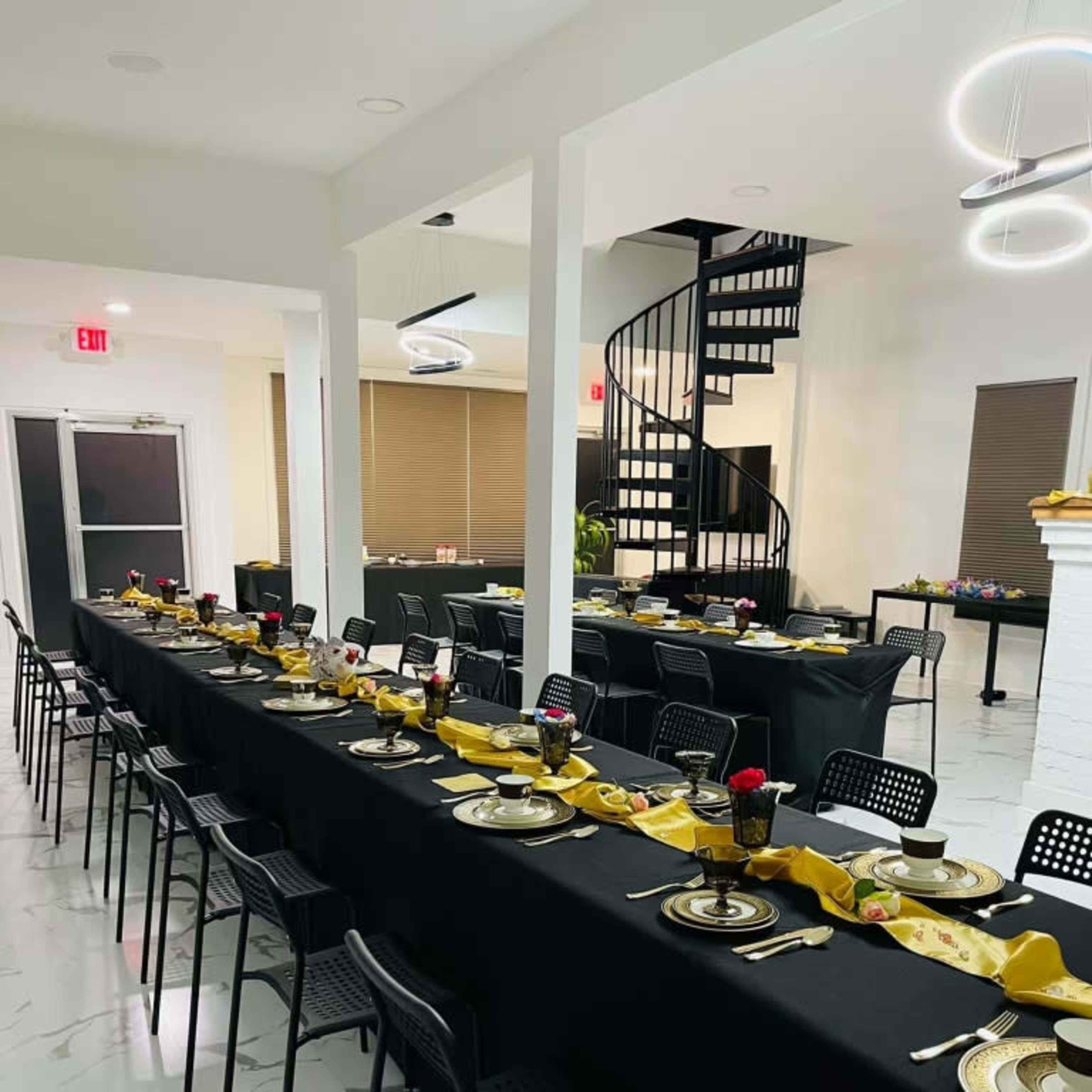 A large dining room with several long tables set with black tablecloths, plates, and floral centerpieces, and a spiral staircase visible in the background.
