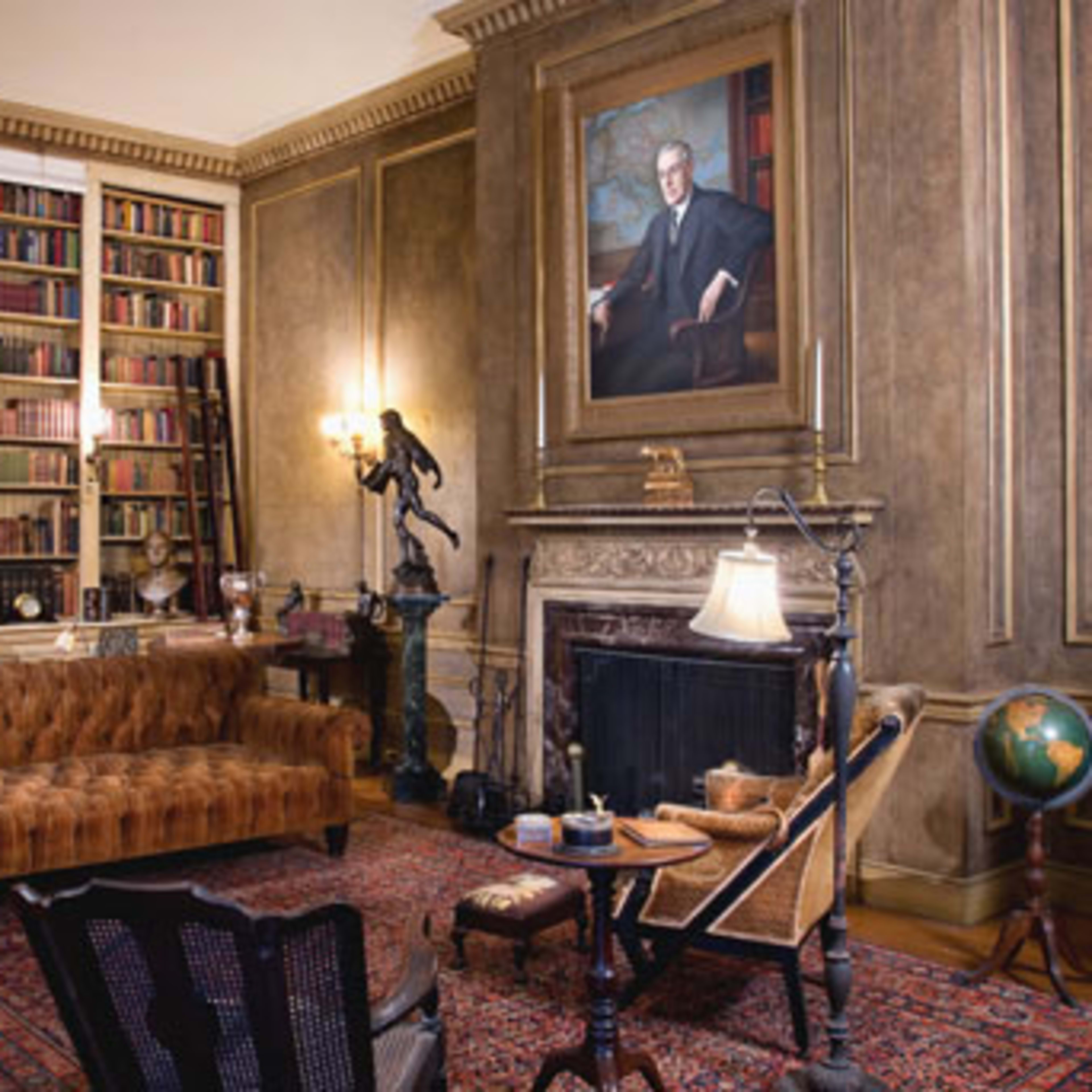 A well-furnished library with a leather sofa, bookshelves filled with books, a portrait on the wall, and a fireplace.
