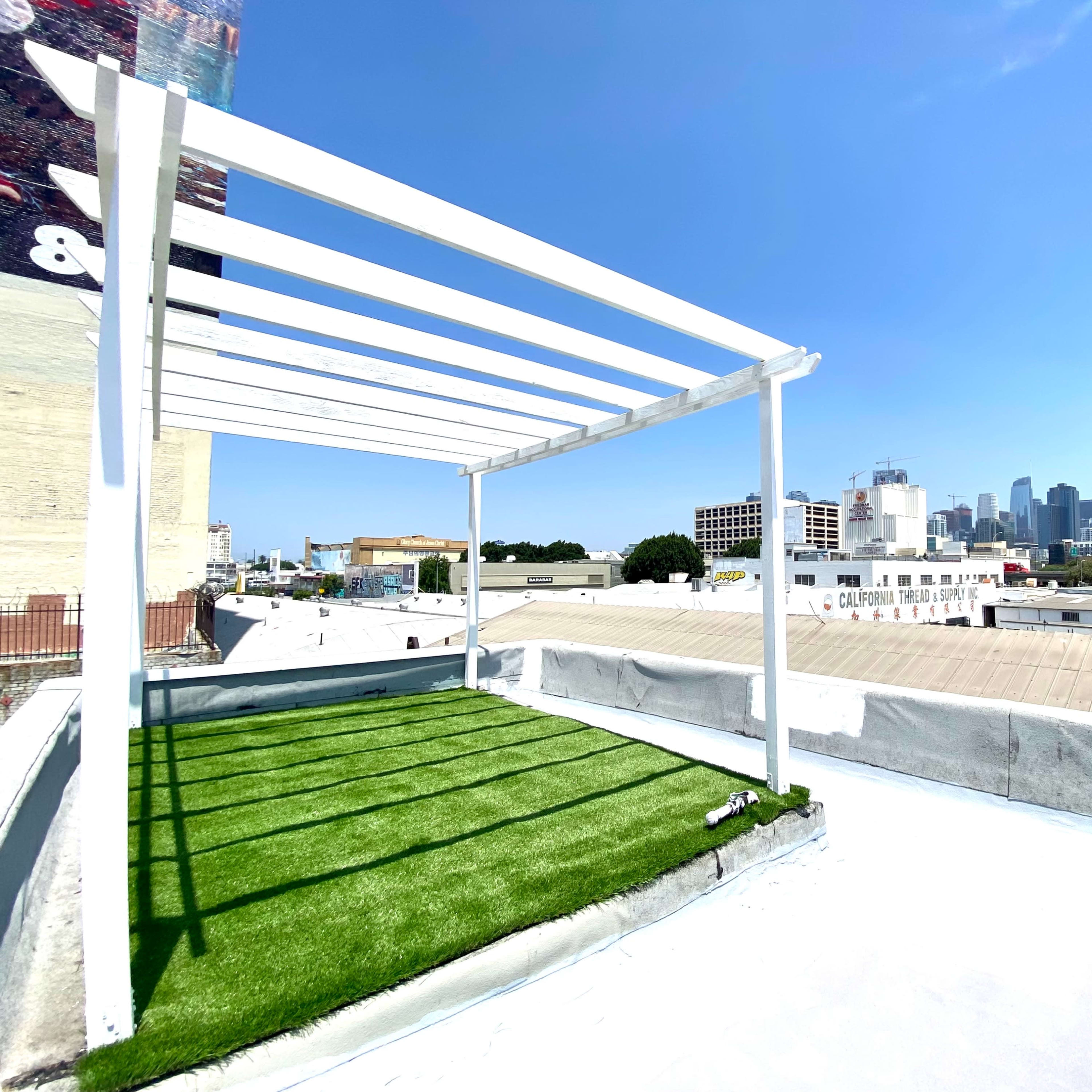 A rooftop terrace with artificial grass and a white pergola overlooks a city skyline under a clear blue sky.