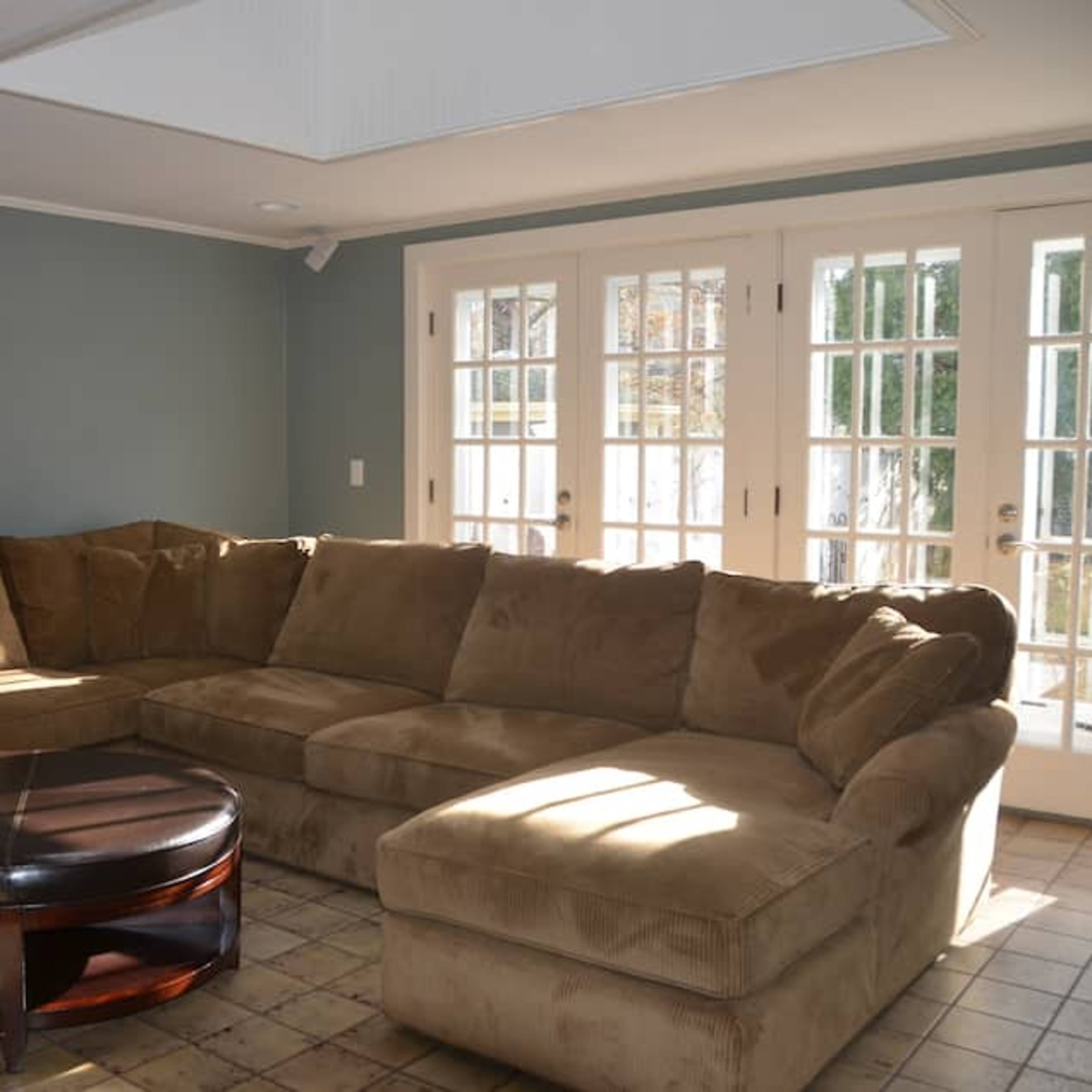 A spacious living room with a large, brown sectional sofa and a round coffee table, illuminated by natural light from double French doors.