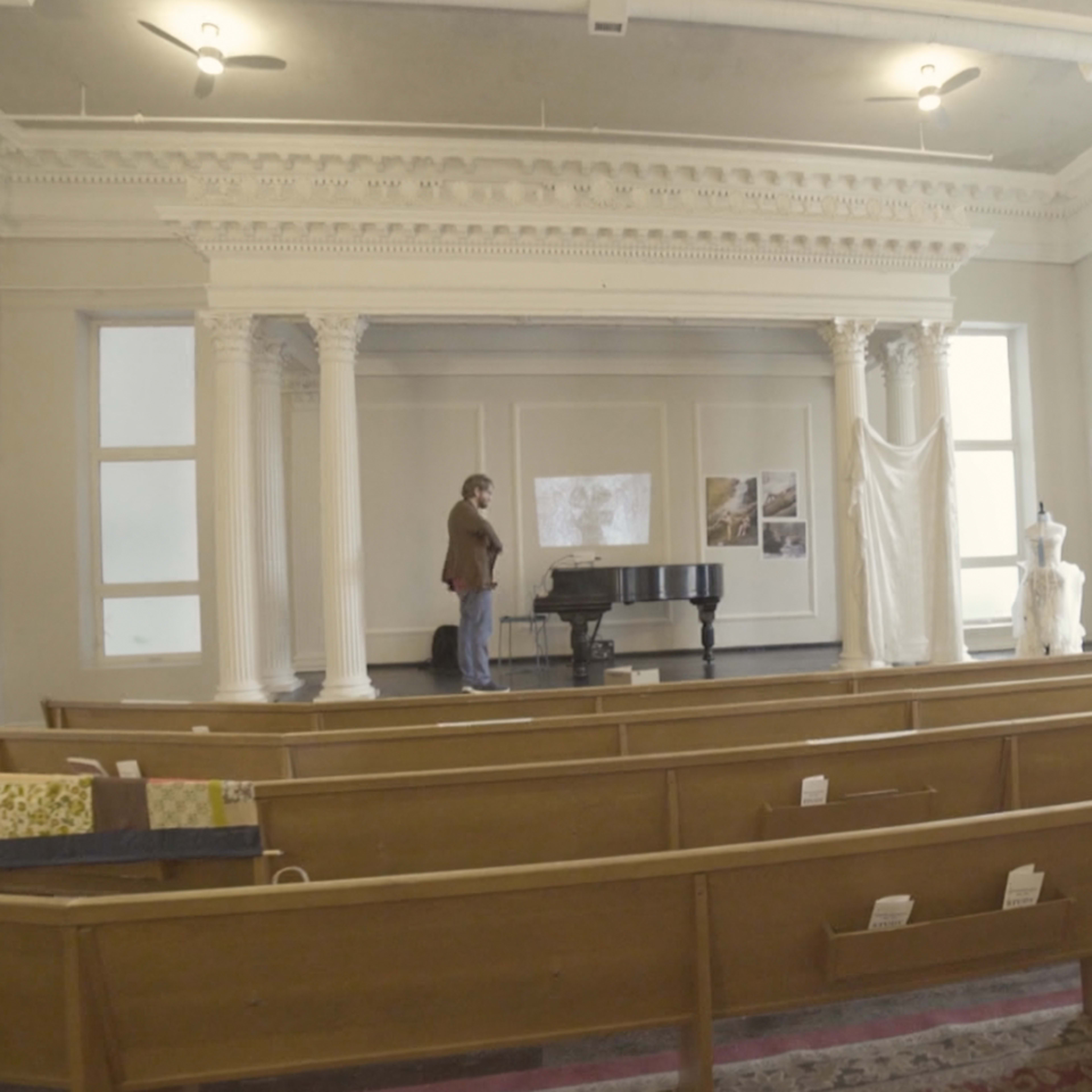 A person stands next to a grand piano on a raised stage surrounded by wooden pews in a spacious room with large windows and high ceilings.