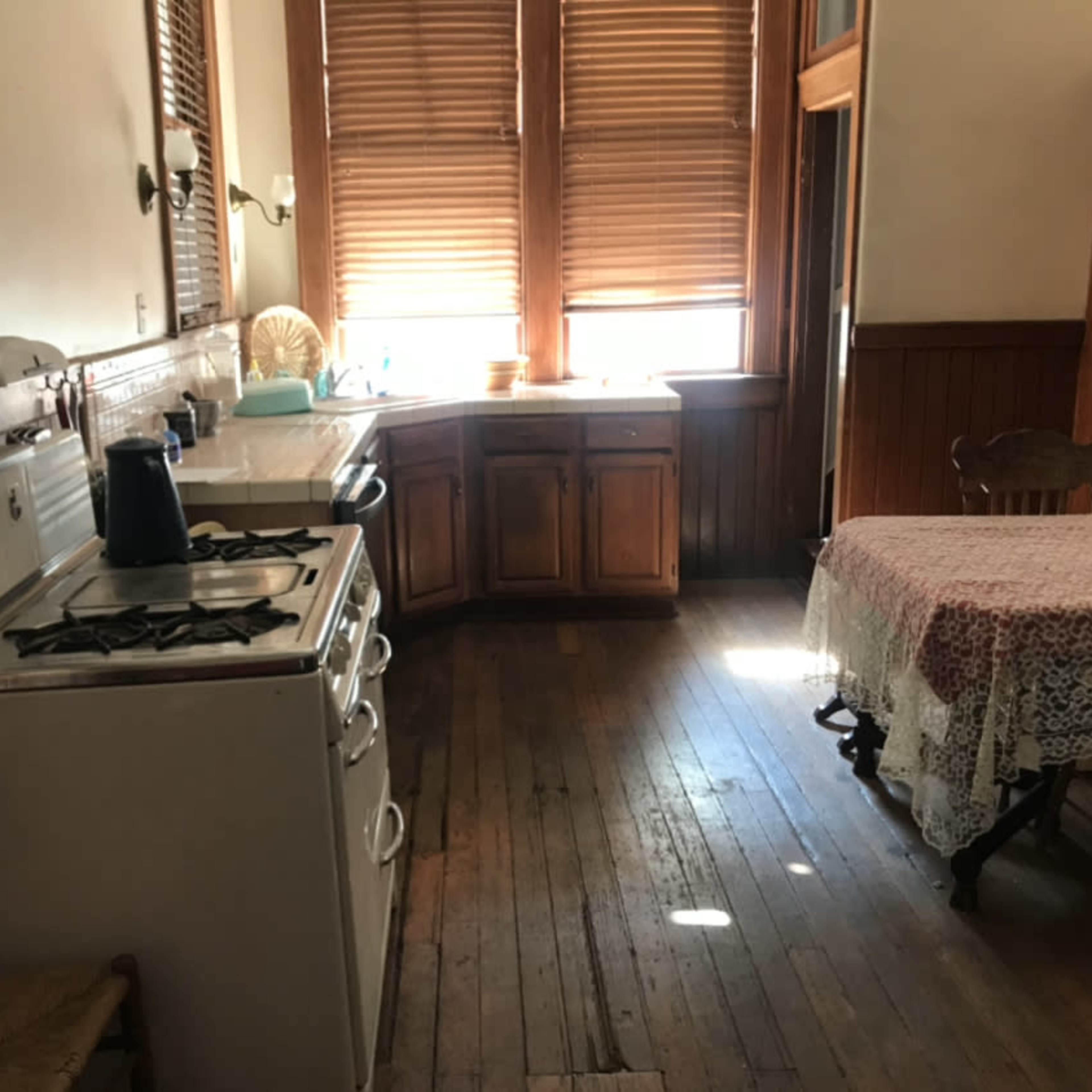 A vintage kitchen with wooden cabinets, a gas stove, and a dining table covered with a lace tablecloth near a window with wooden blinds.