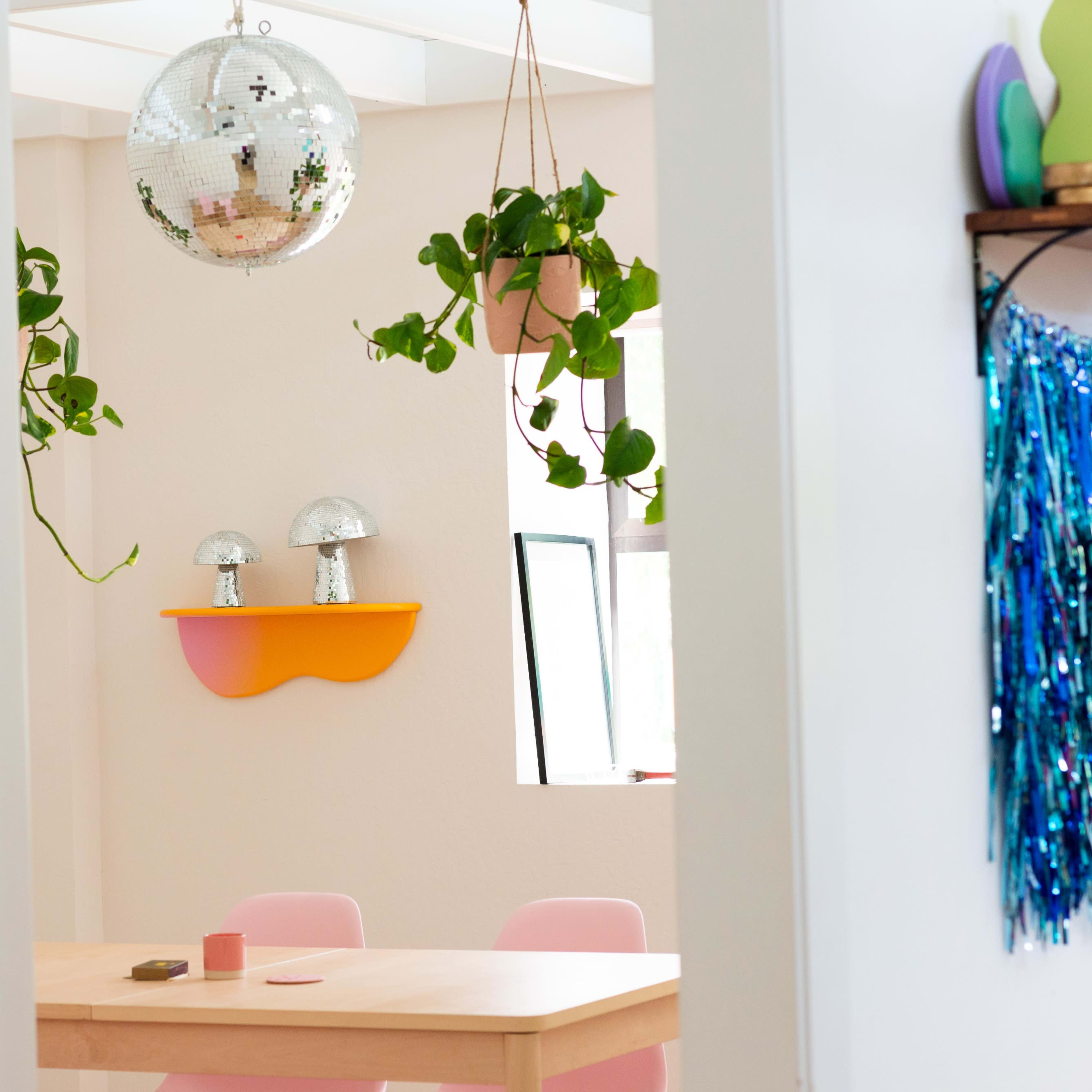 A bright dining area with a wooden table, pink chairs, and a disco ball hanging from the ceiling, alongside potted plants on shelves.