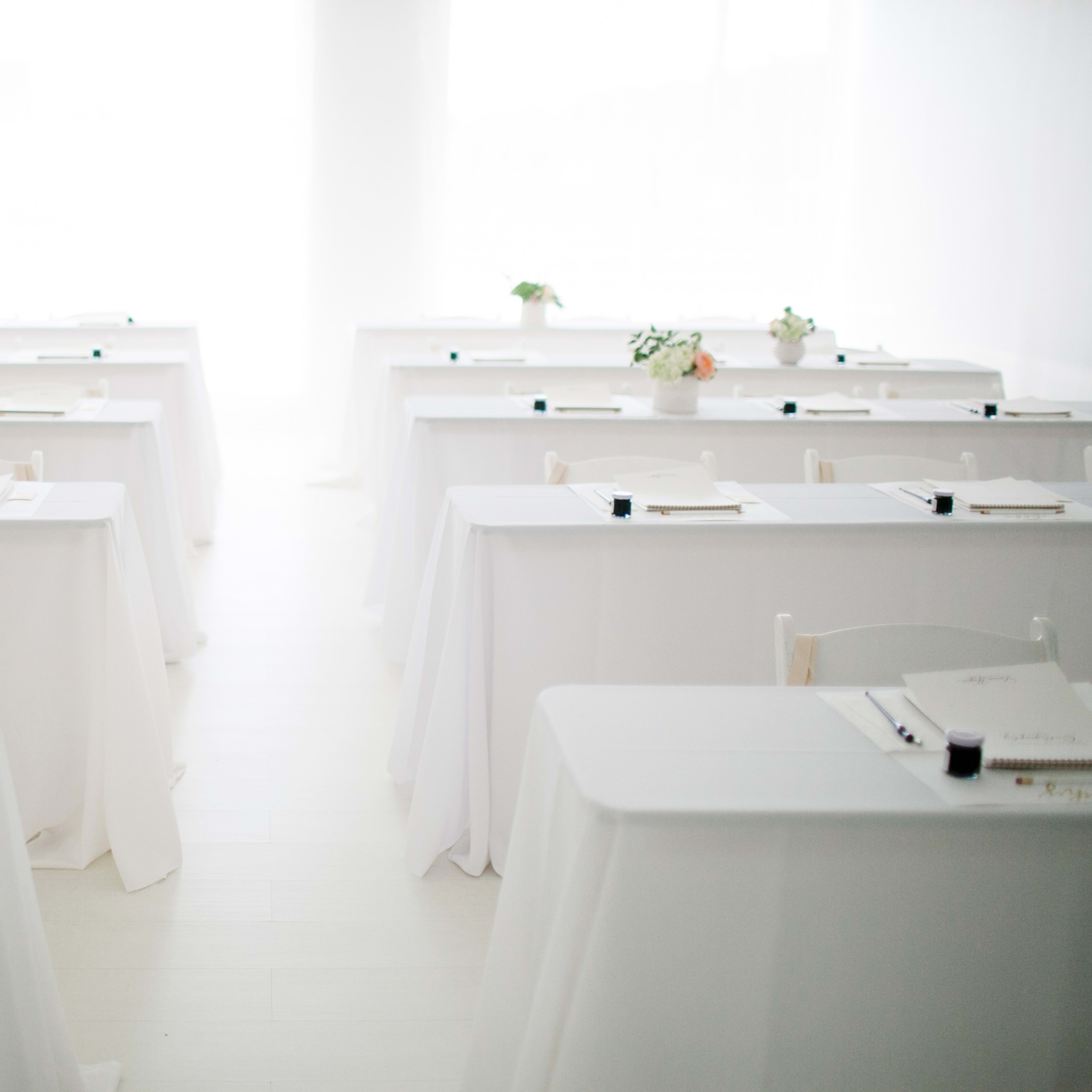 A bright, minimalistic room set up for a seminar or workshop, featuring neatly arranged tables with white tablecloths and floral centerpieces.
