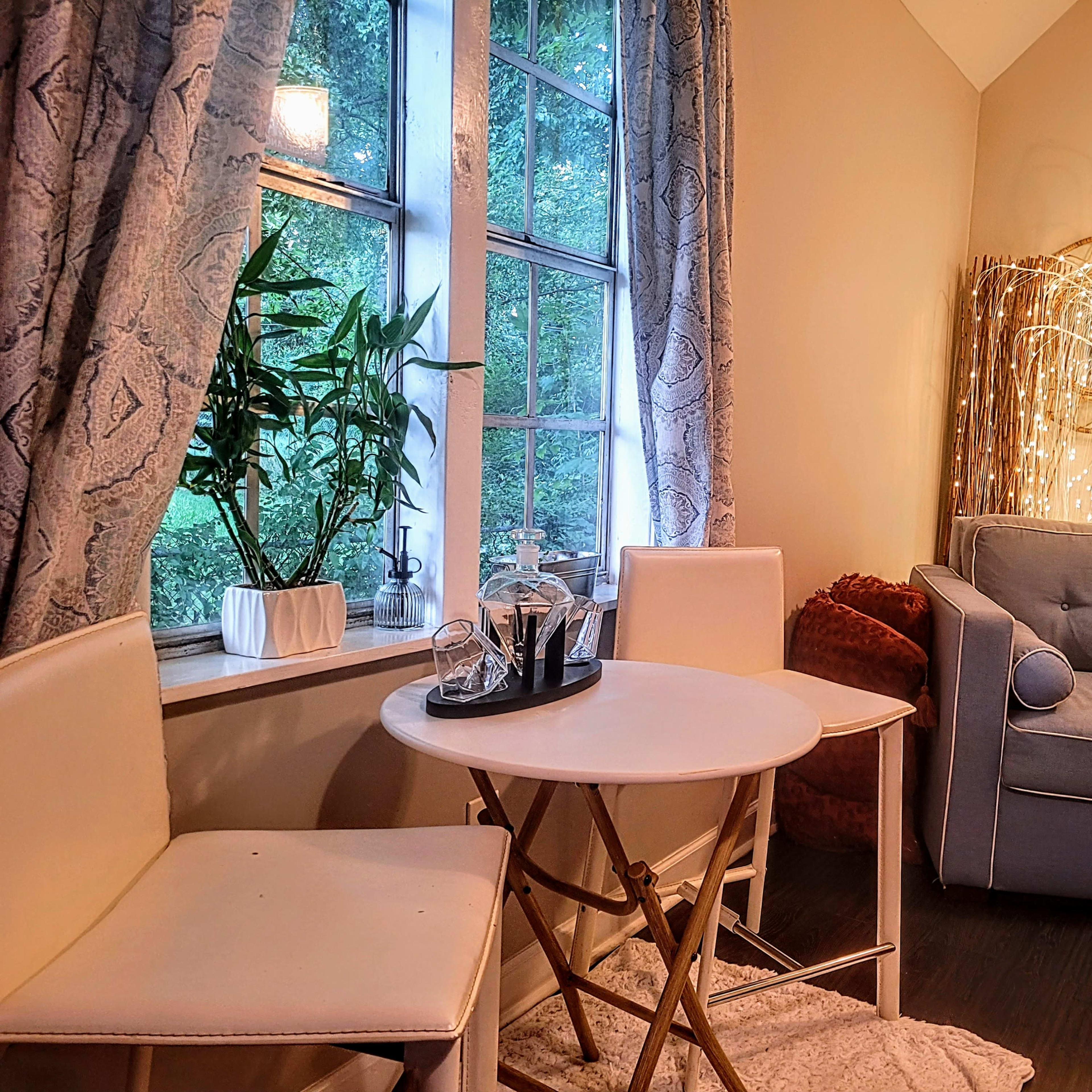 A small dining area features a round table with two chairs beside a window, a cozy couch, and decorative lighting in the background.