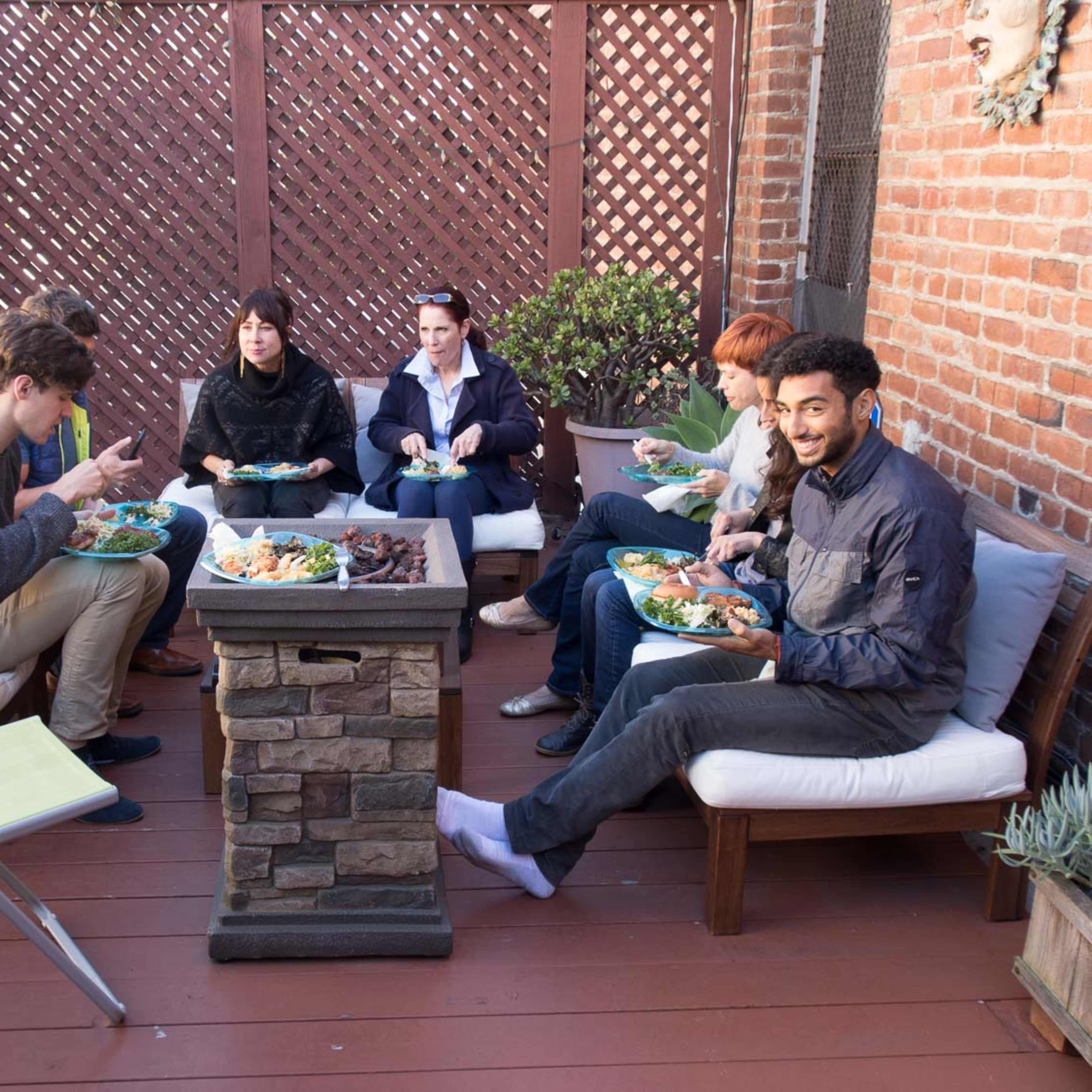 A group of six people sit around a stone fire pit on a patio, enjoying a meal together.