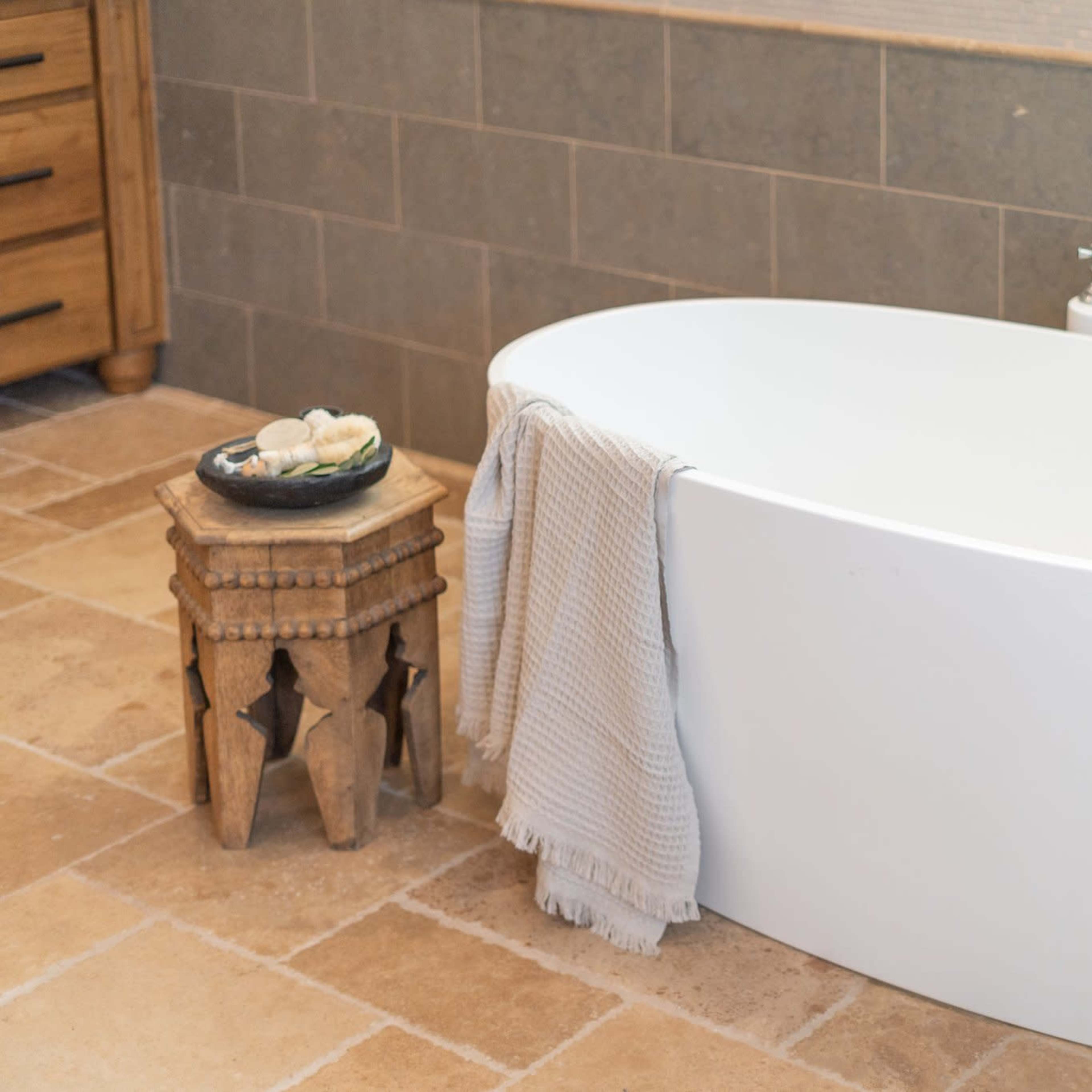 A modern, freestanding white bathtub next to a wooden stool adorned with decorative stones and a light-colored towel.