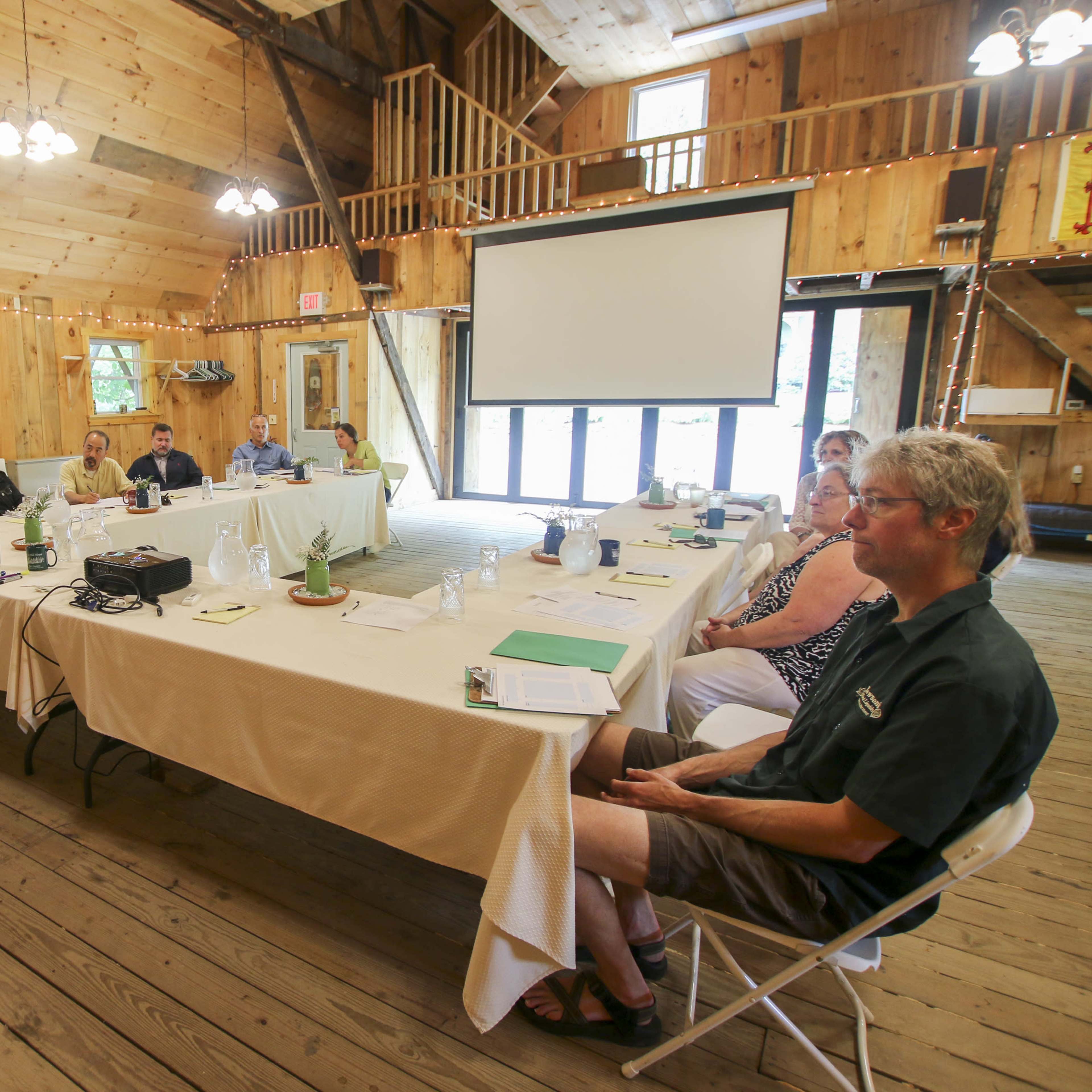 A group of people is seated at a long table in a wooden meeting room while a woman stands and speaks at the front.