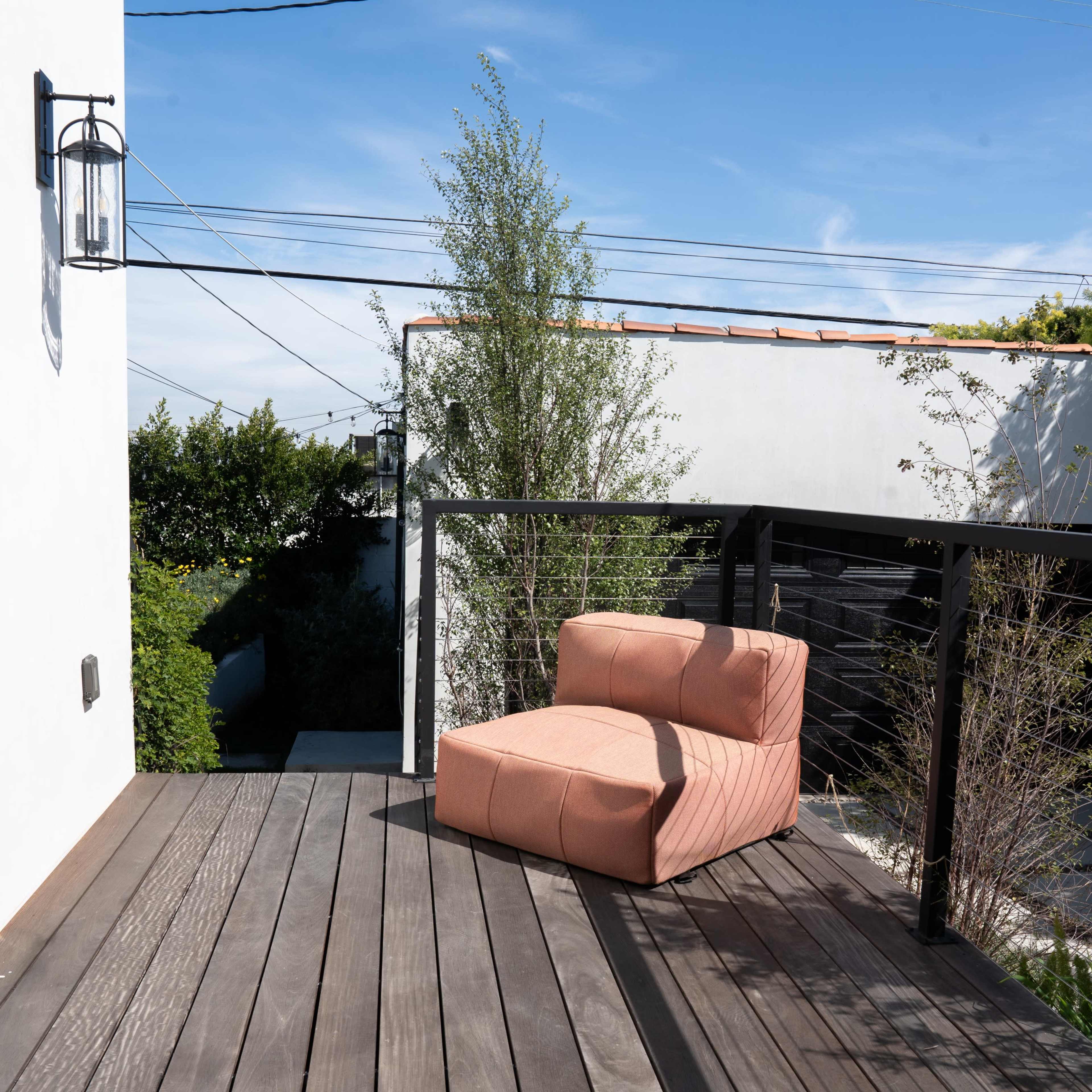 A single coral-colored lounge chair sits on a wooden deck surrounded by greenery under a clear blue sky.
