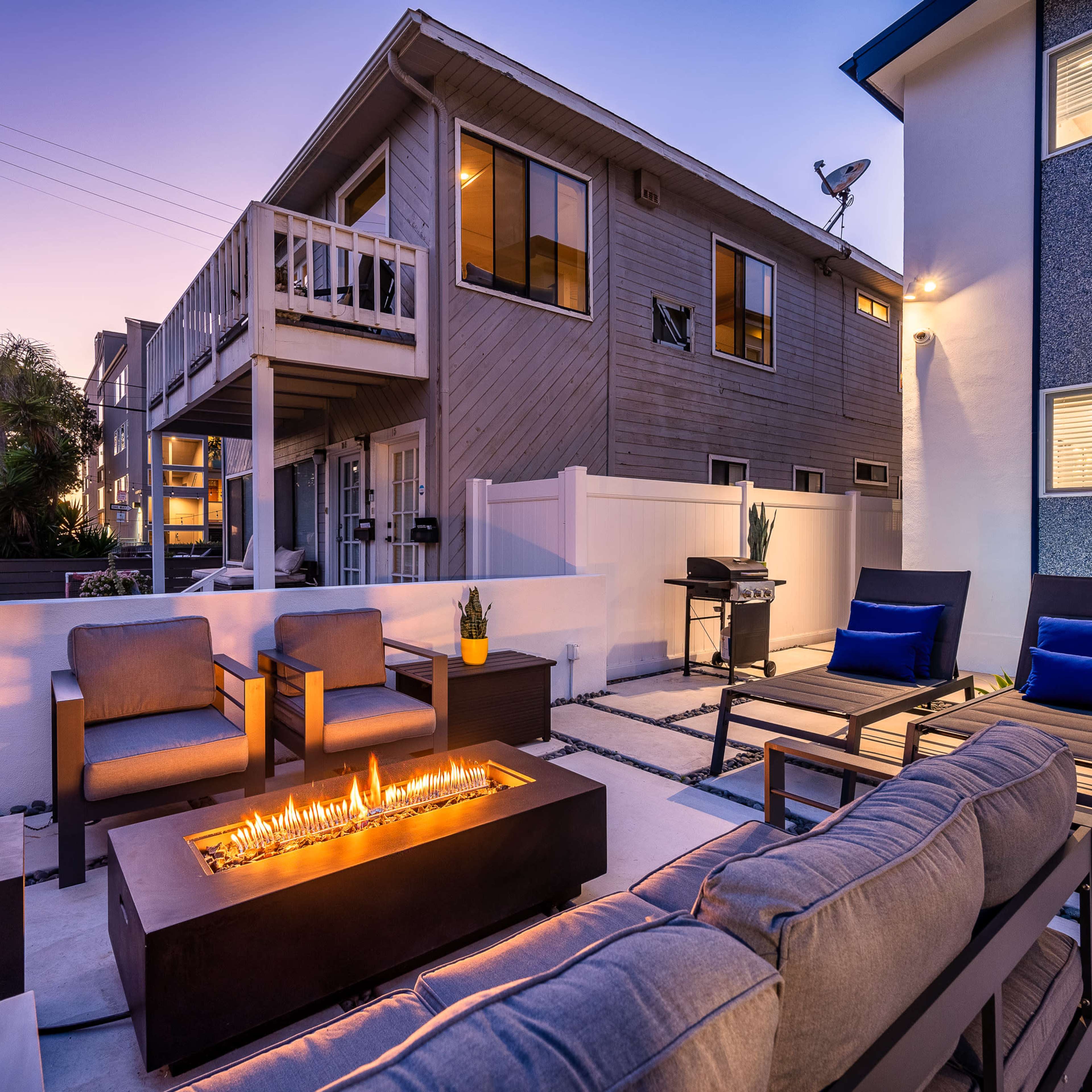 A modern outdoor seating area features a fire pit surrounded by chairs and a sofa, with a building in the background illuminated by twilight.